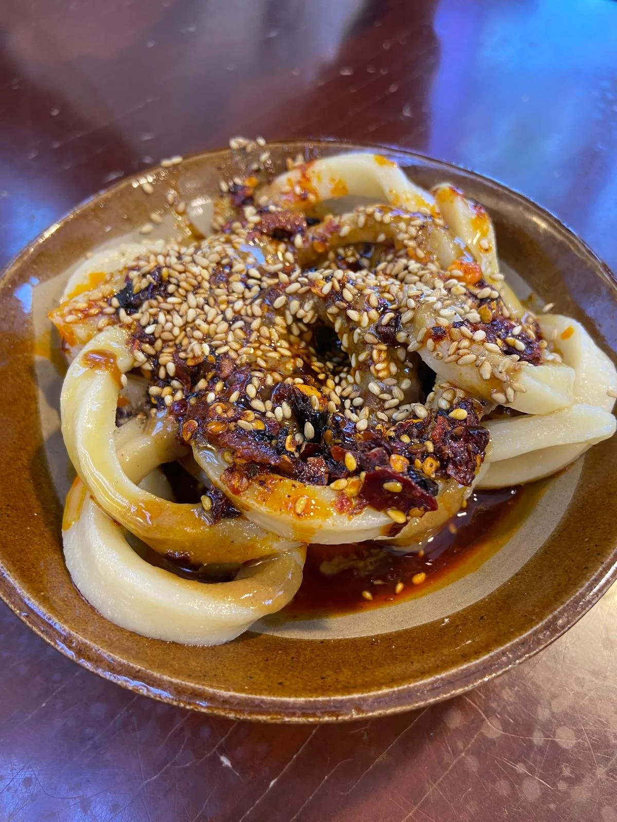 A bowl of pasta topped with chili flakes and sesame seeds, served on a brown dish on a dark wooden table.