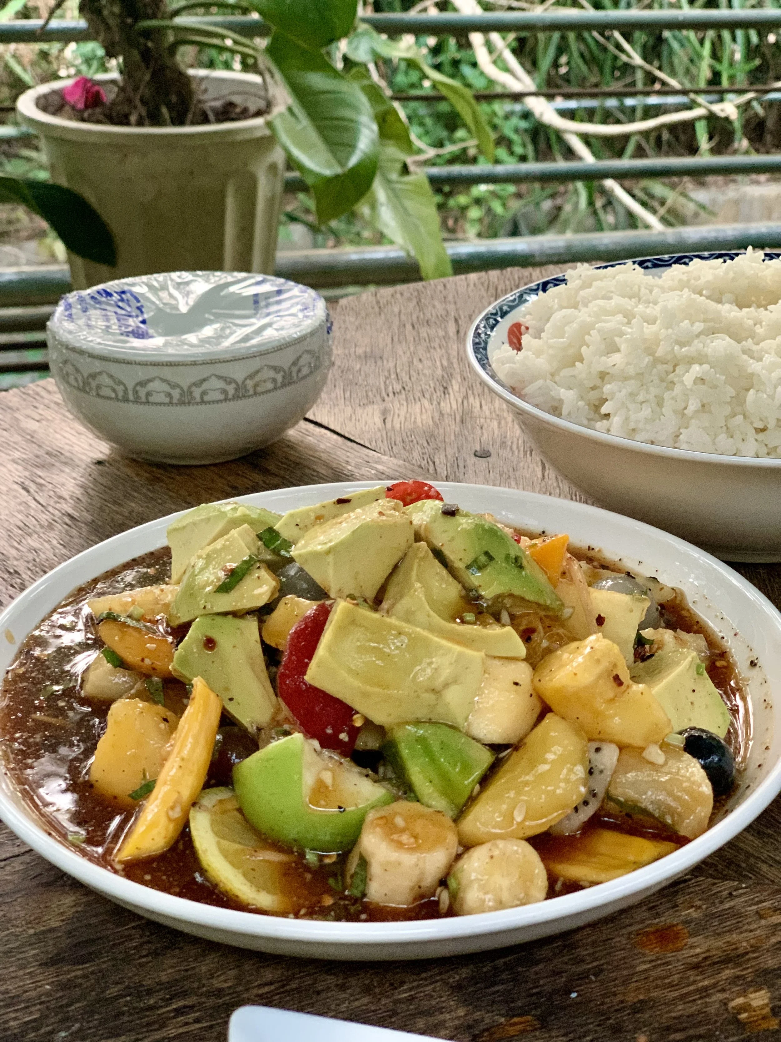 A plate of vegetable stir-fry with rice, set on a wooden table outdoors, with a small bowl and a bowl of white rice in the background, and potted plants on a balcony railing.