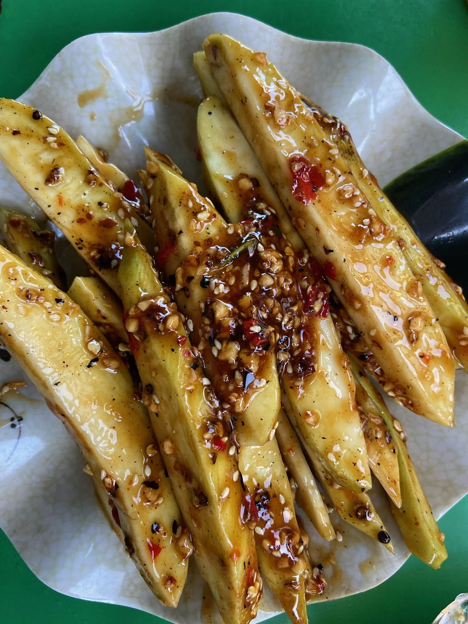 Cooked sliced eggplant with sauce, sesame seeds, and red chili flakes on white paper, served on a green surface.