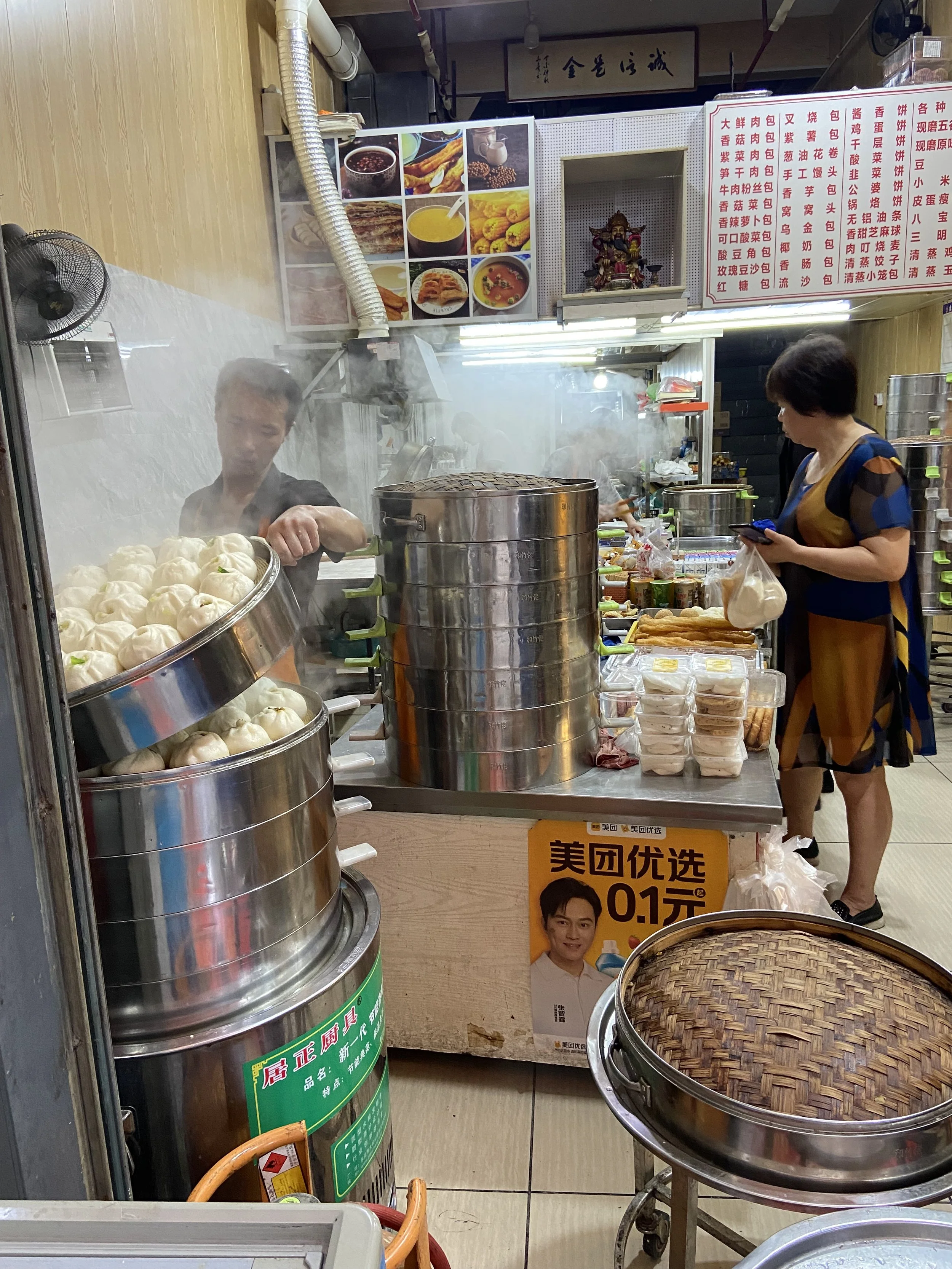 A busy Chinese food stall with steam rising, steaming bamboo baskets filled with dumplings, a woman looking at her phone, and various dishes and ingredients on the counter.