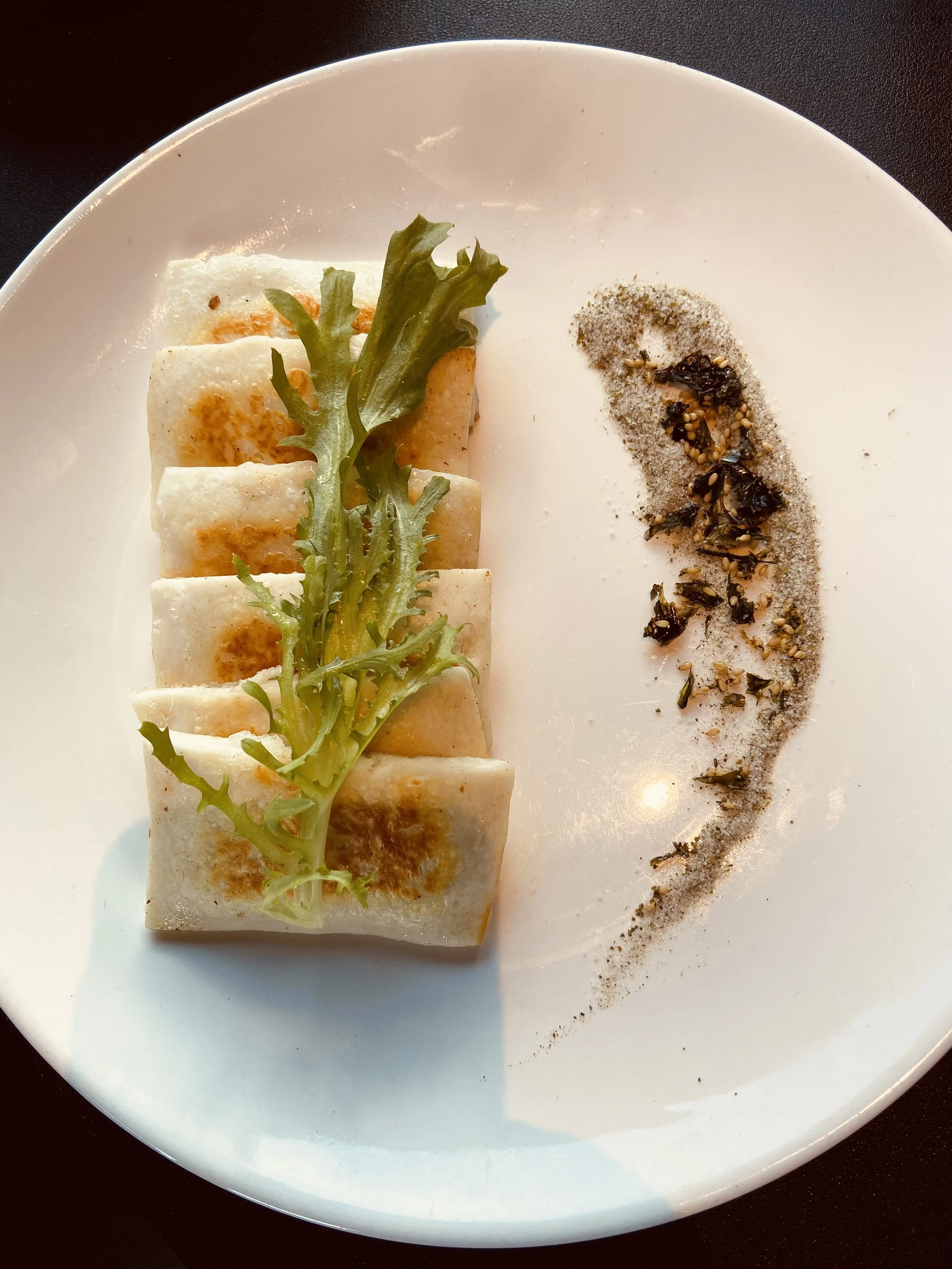 Six pan-fried spring rolls topped with green lettuce on a white plate, with black pepper, sesame seeds, and chili flakes garnished with salt on the side.