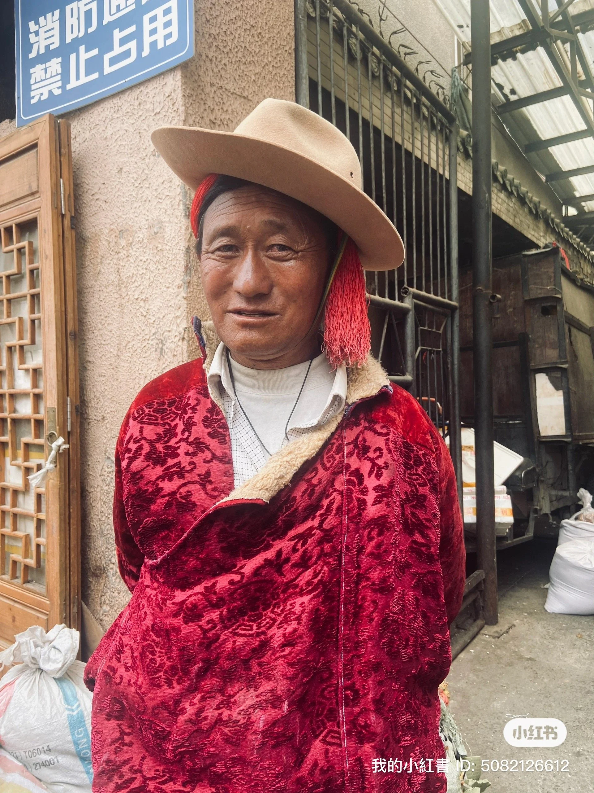 An elderly man in traditional attire, wearing a red floral-patterned cloak and a beige wide-brimmed hat, stands outdoors in front of a building with Chinese signs. He has a slight smile and a red cloth tied around his head.