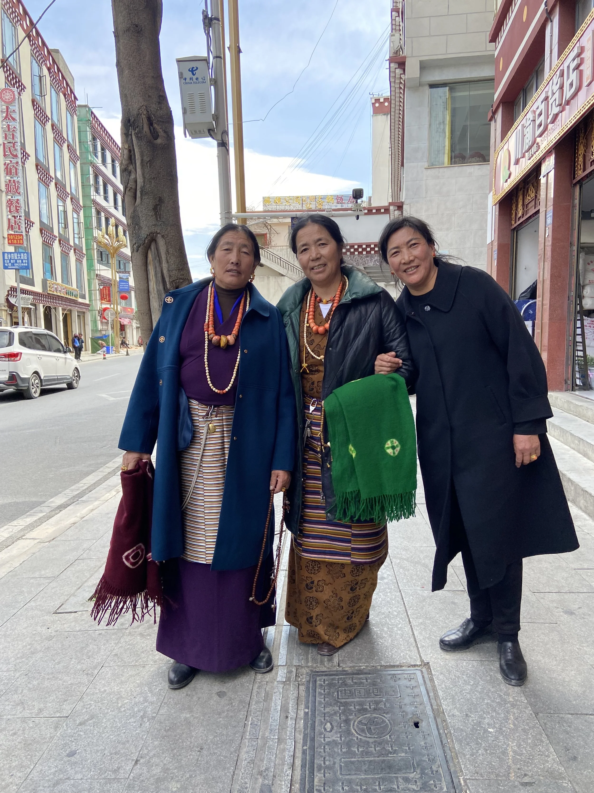 Three women standing on a city sidewalk, dressed in traditional clothing with colorful necklaces and shawls, posing for a photo.