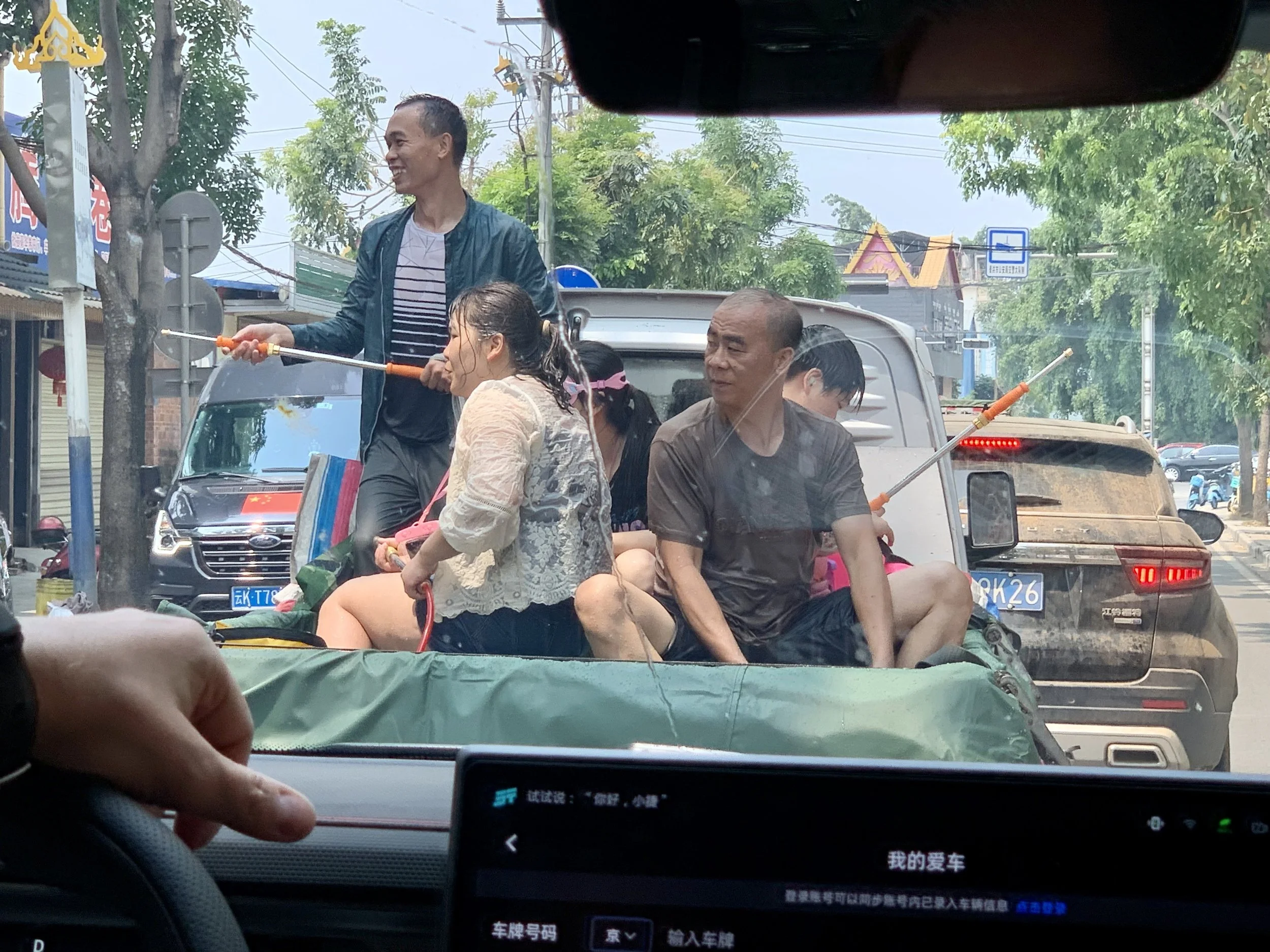 A group of people riding in the back of a small open truck, with some individuals holding rod-like objects, on a city street lined with trees and buildings, viewed from inside a car.