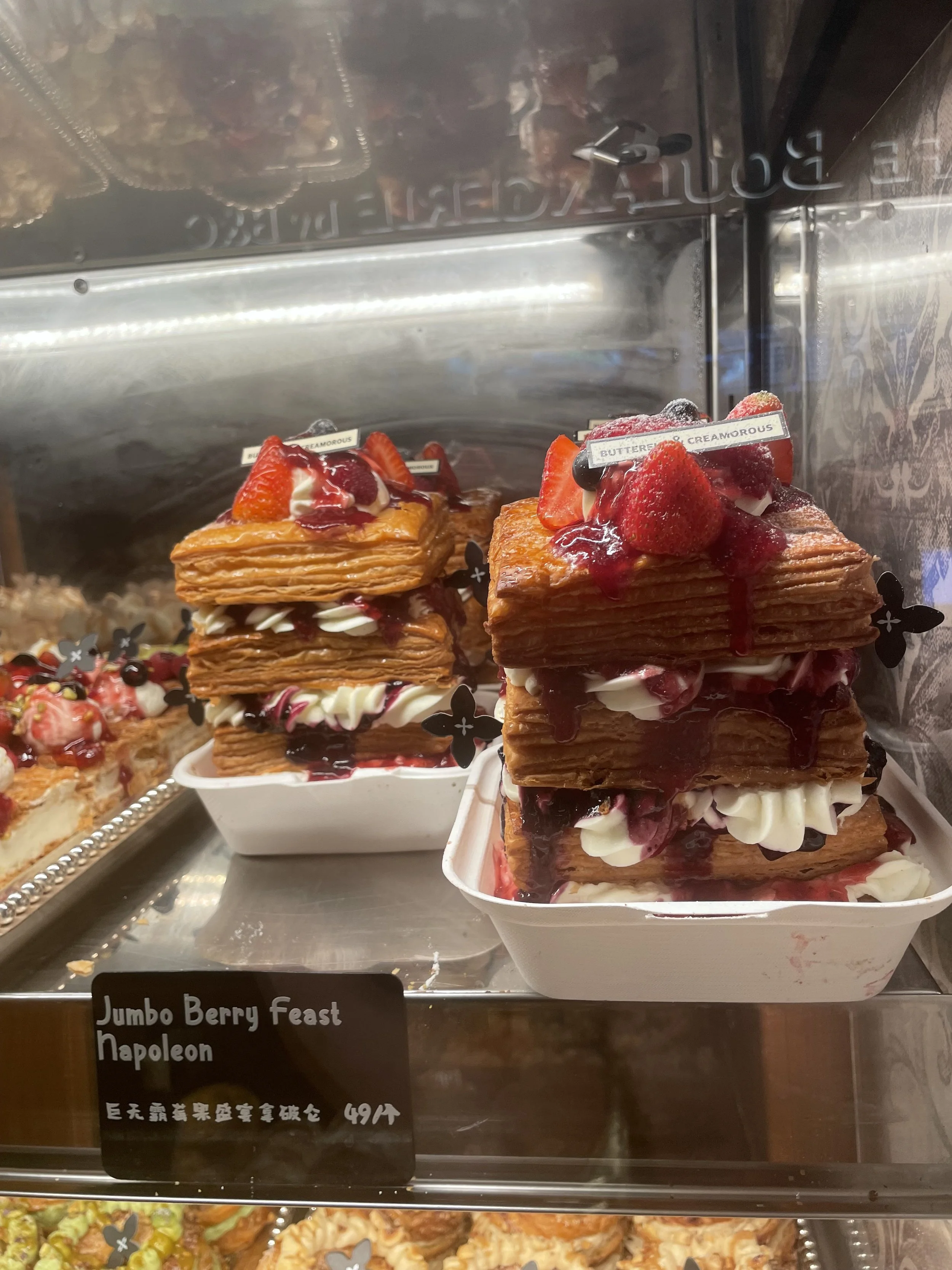 Display of layered pastry desserts topped with strawberries, drizzled with berry sauce, and decorated with whipped cream and small chocolate shavings. The desserts are labeled "Jumbo Berry Feast Napolean."