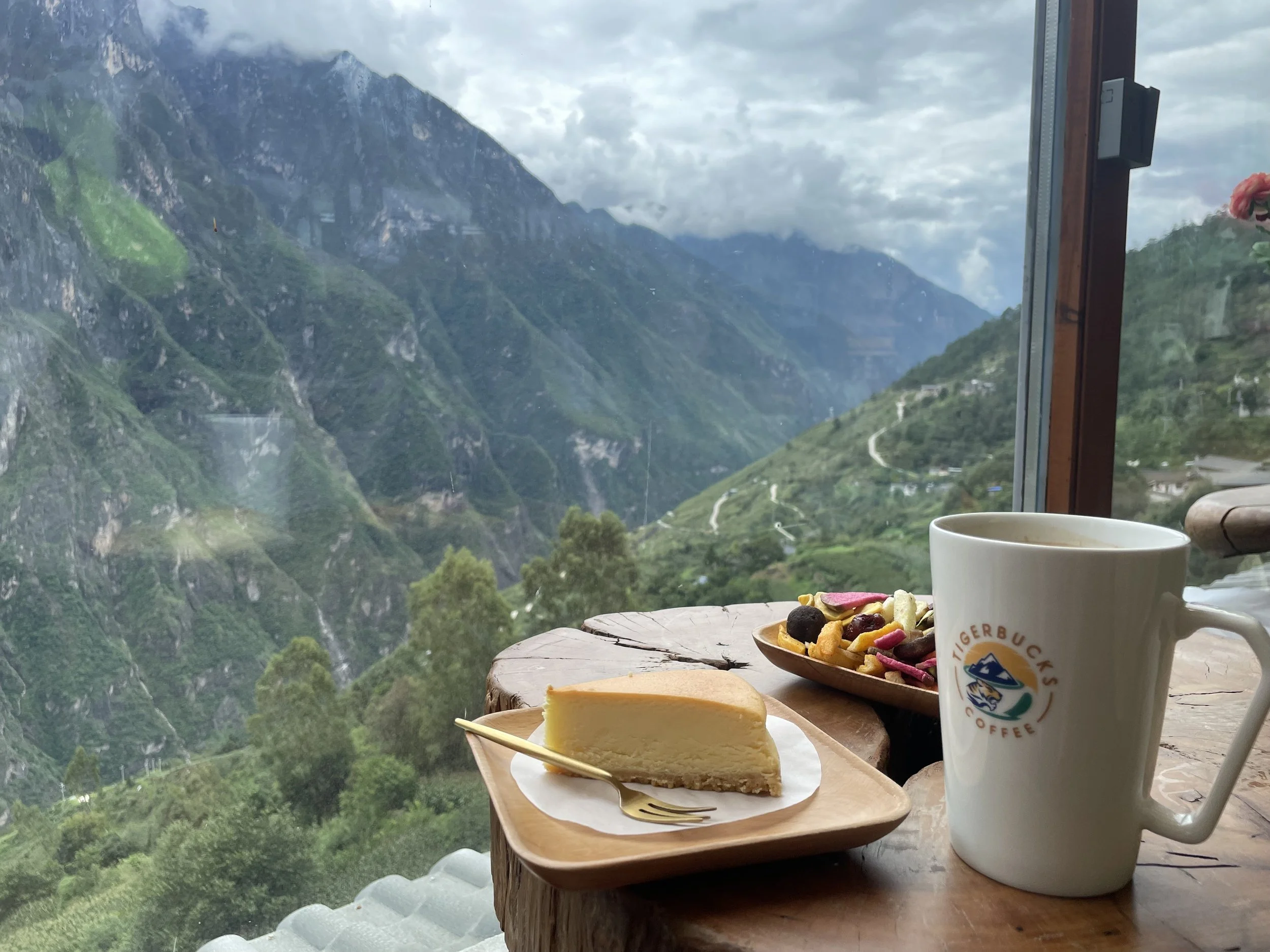 Cheesecake, trail mix, and coffee on a wooden table with a mountain view through a window