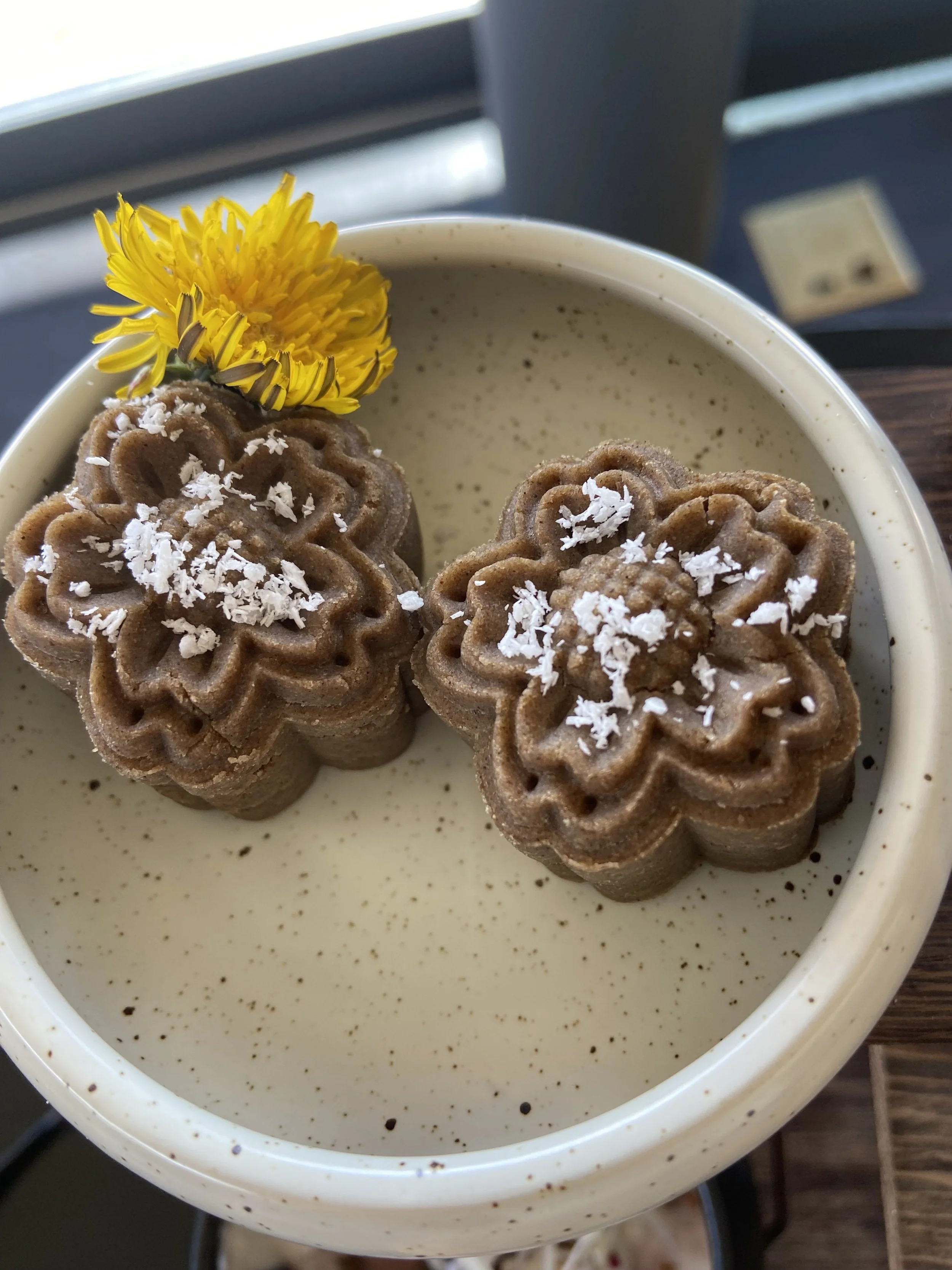 Two flower-shaped chocolate cookies with shredded coconut on top, garnished with a yellow dandelion flower, in a cream-colored bowl.