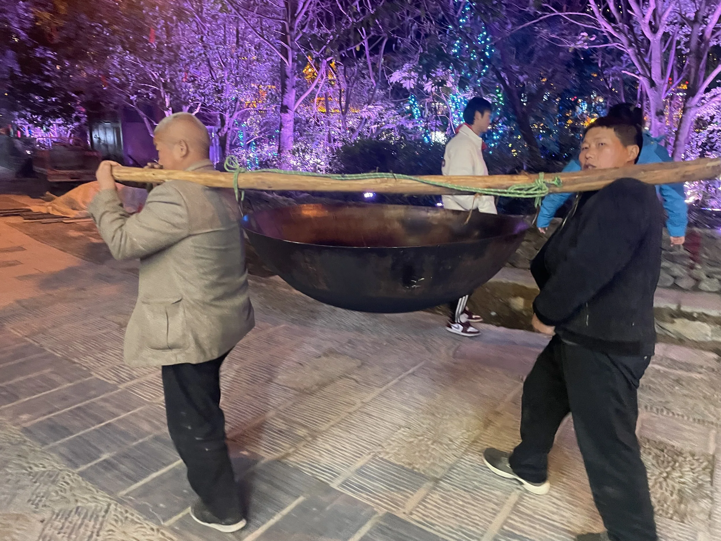 Two men carrying a large black wok on a wooden pole at night, with colorful lights illuminating trees in the background.