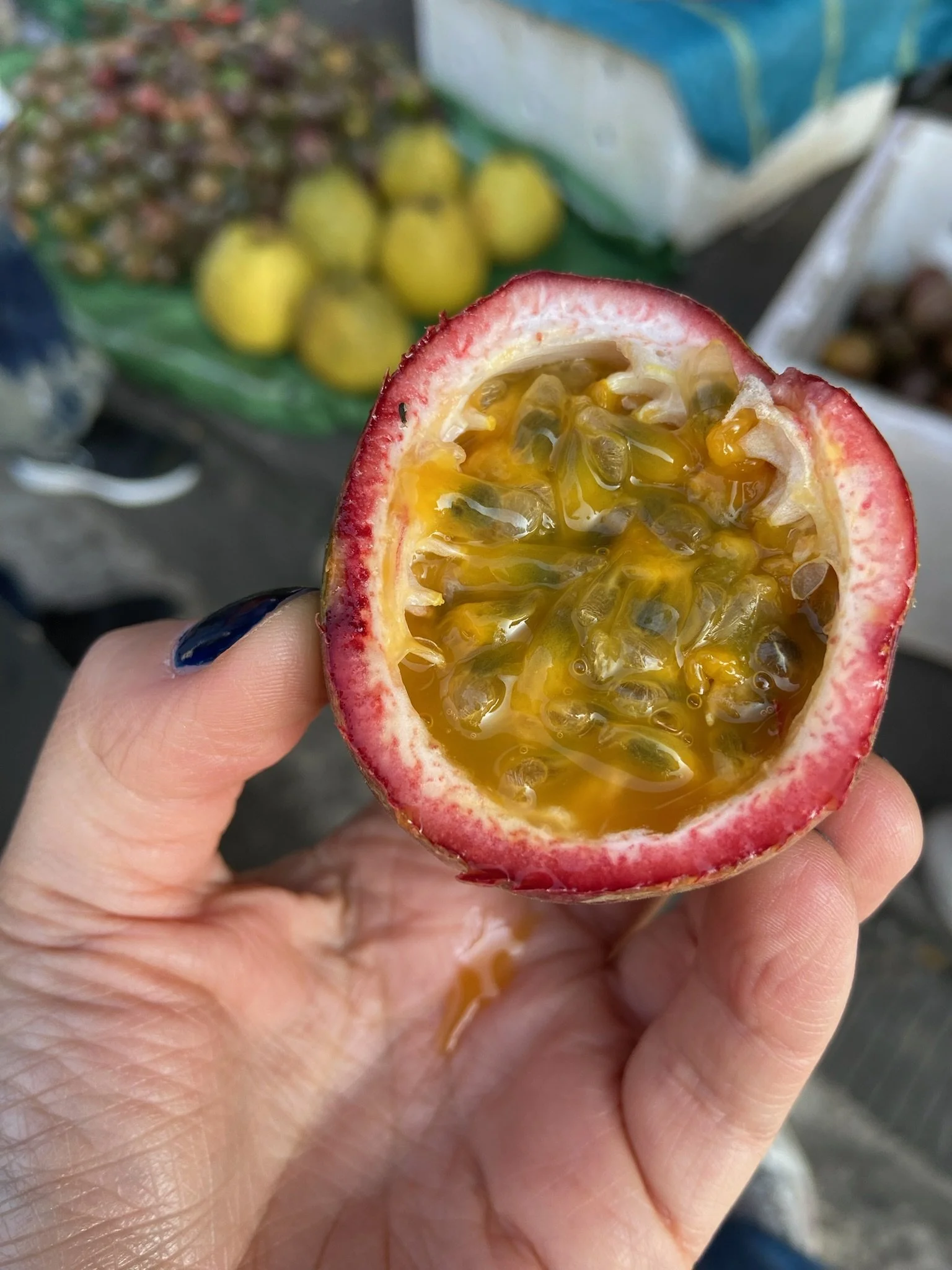 Close-up of a person's hand holding a halved passion fruit with bright yellow seeds inside, with a background of green guava and other produce at a market.
