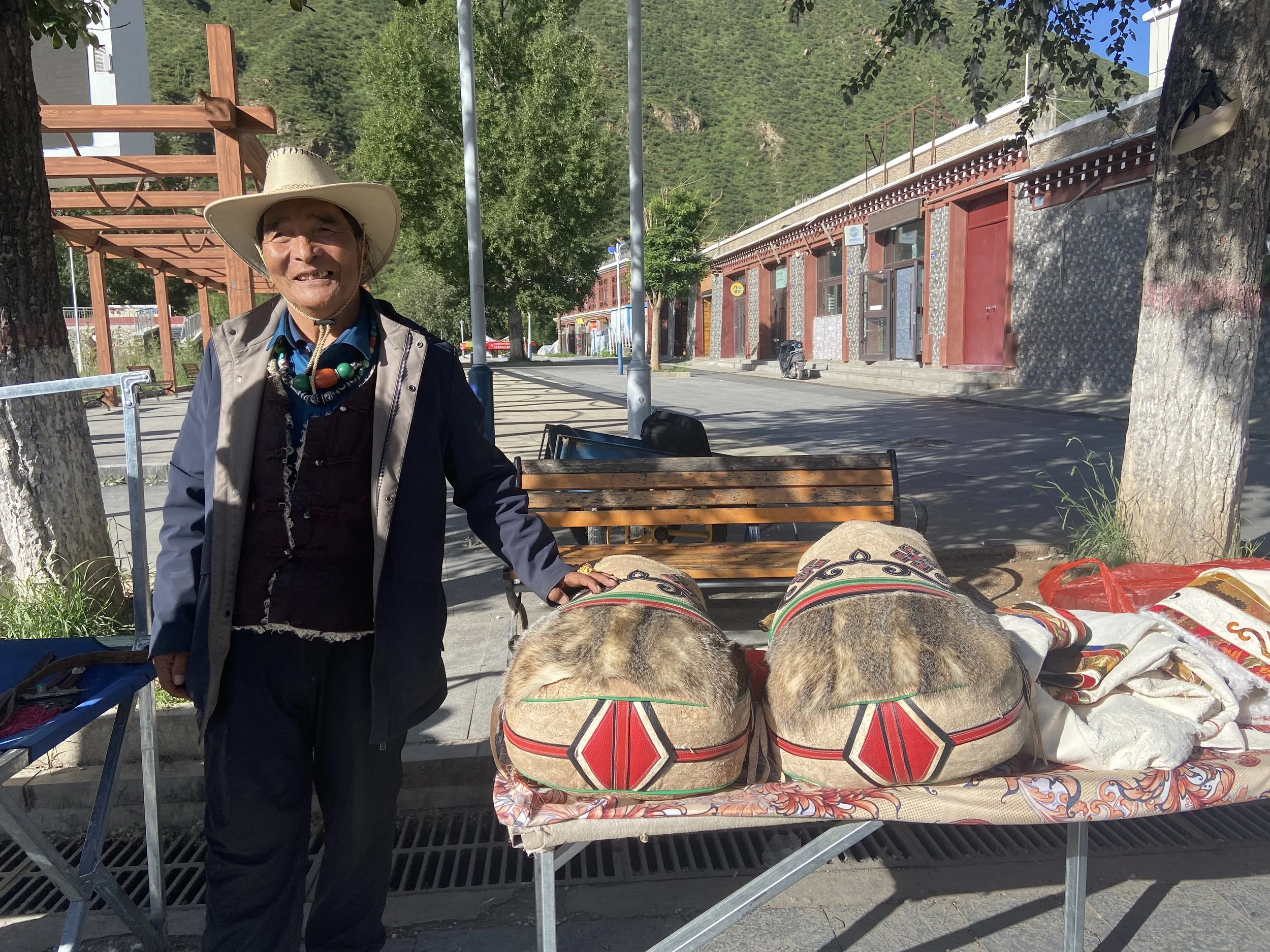 A man in a beige cowboy hat and a dark jacket stands next to a table displaying traditional woven items, with two large, round bundles wrapped in decorated fabric. The scene is outdoors on a sunny day with trees, a building, and mountains in the back
