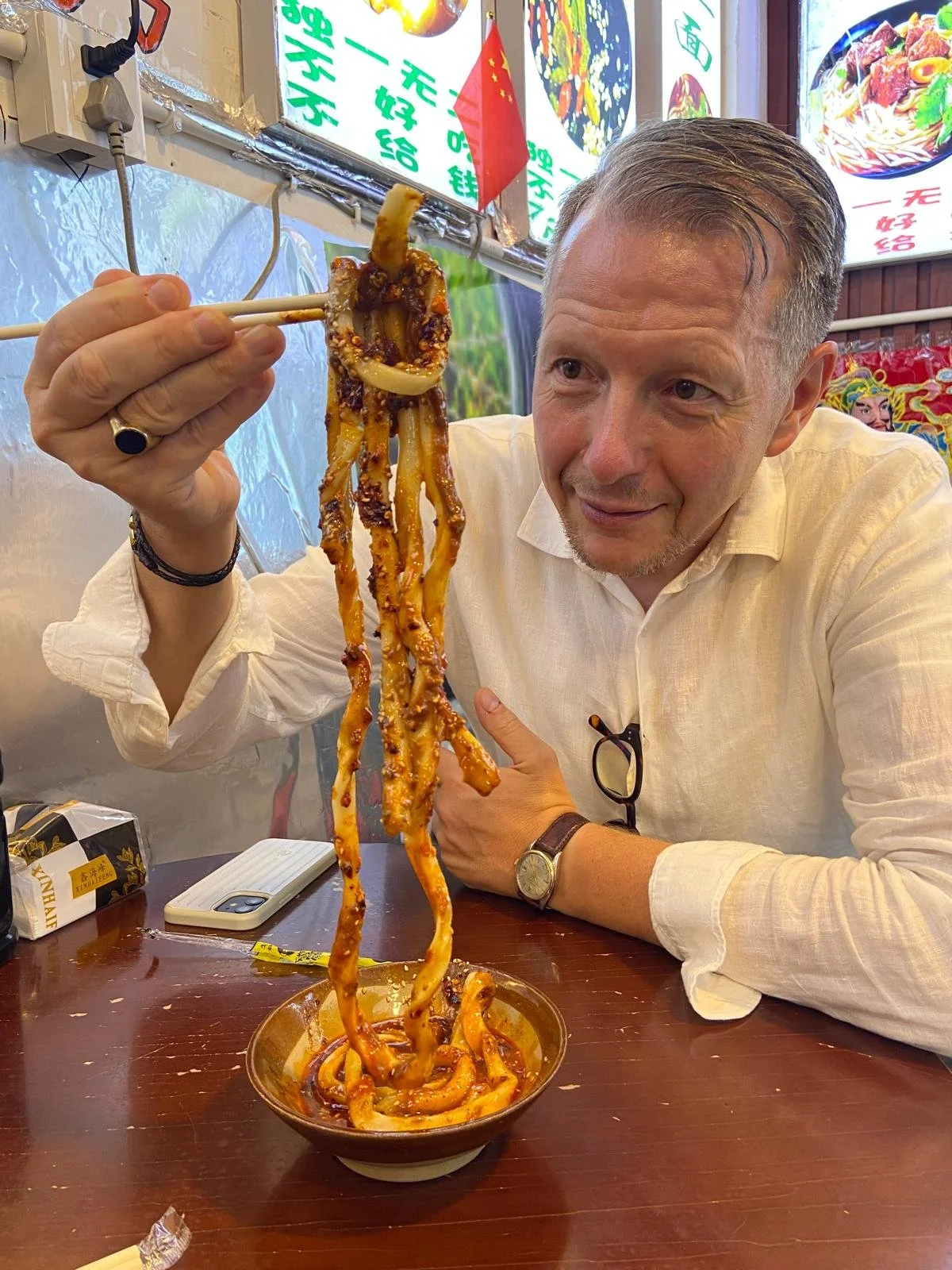 A man in a white shirt eating spicy noodles with chopsticks in a colorful Asian restaurant.