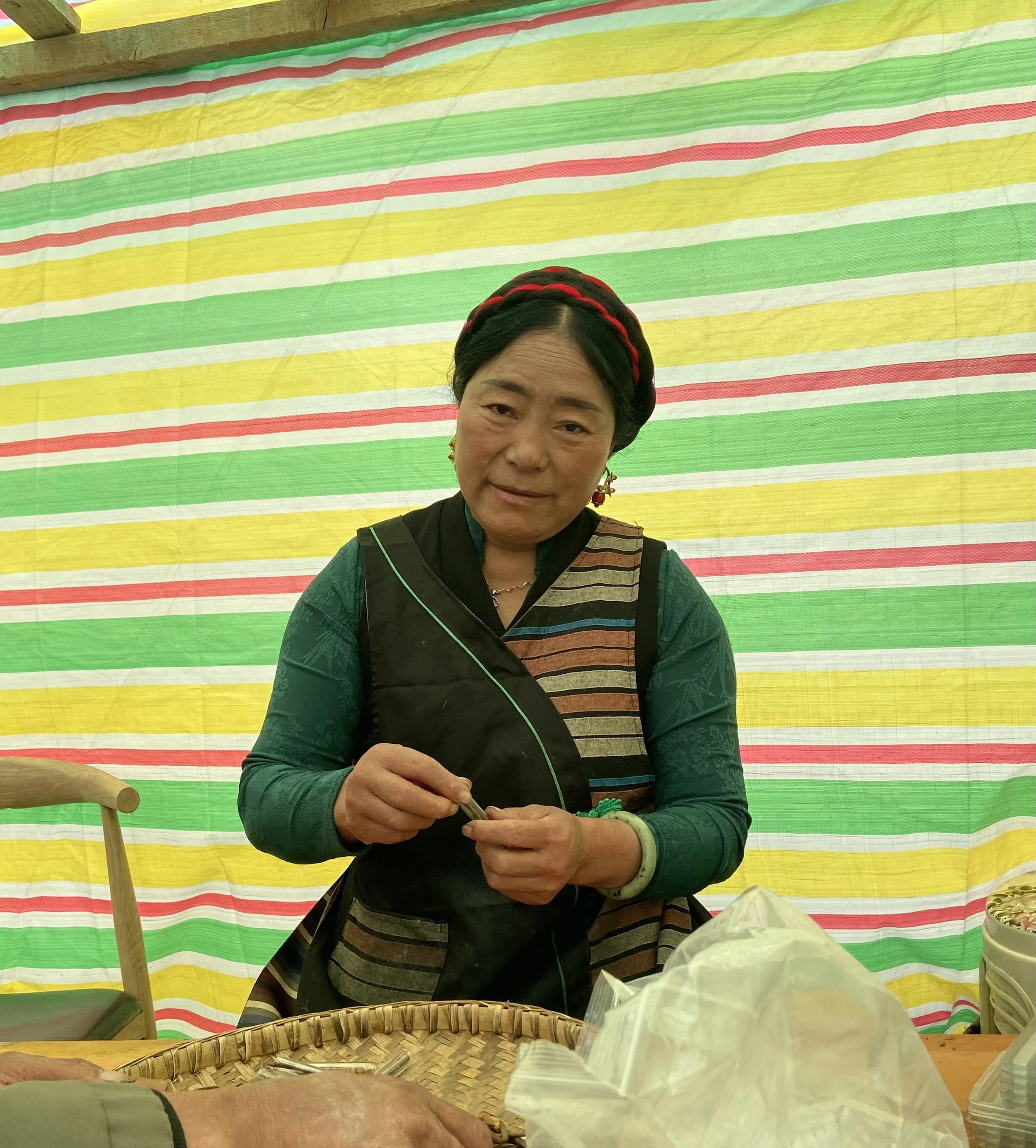 Woman in traditional clothing selling or preparing items at a market stall with a colorful striped background.