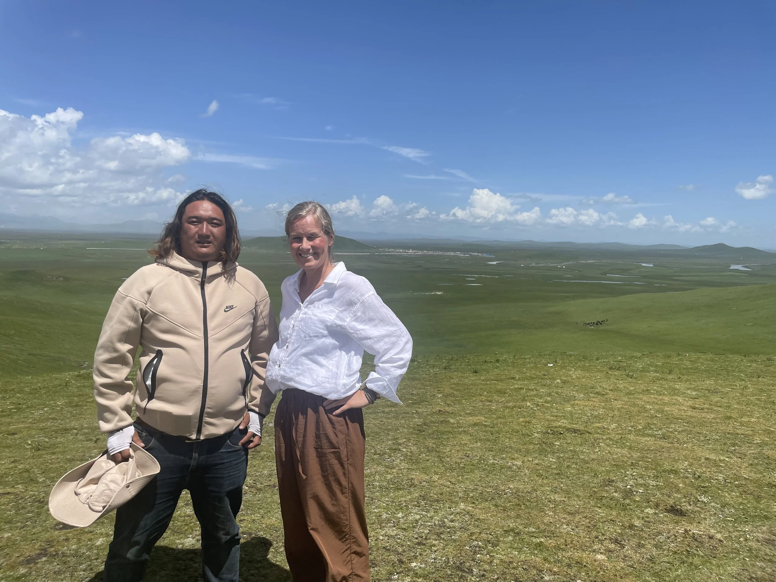 Two people standing outdoors in a vast green landscape with a blue sky and scattered clouds.