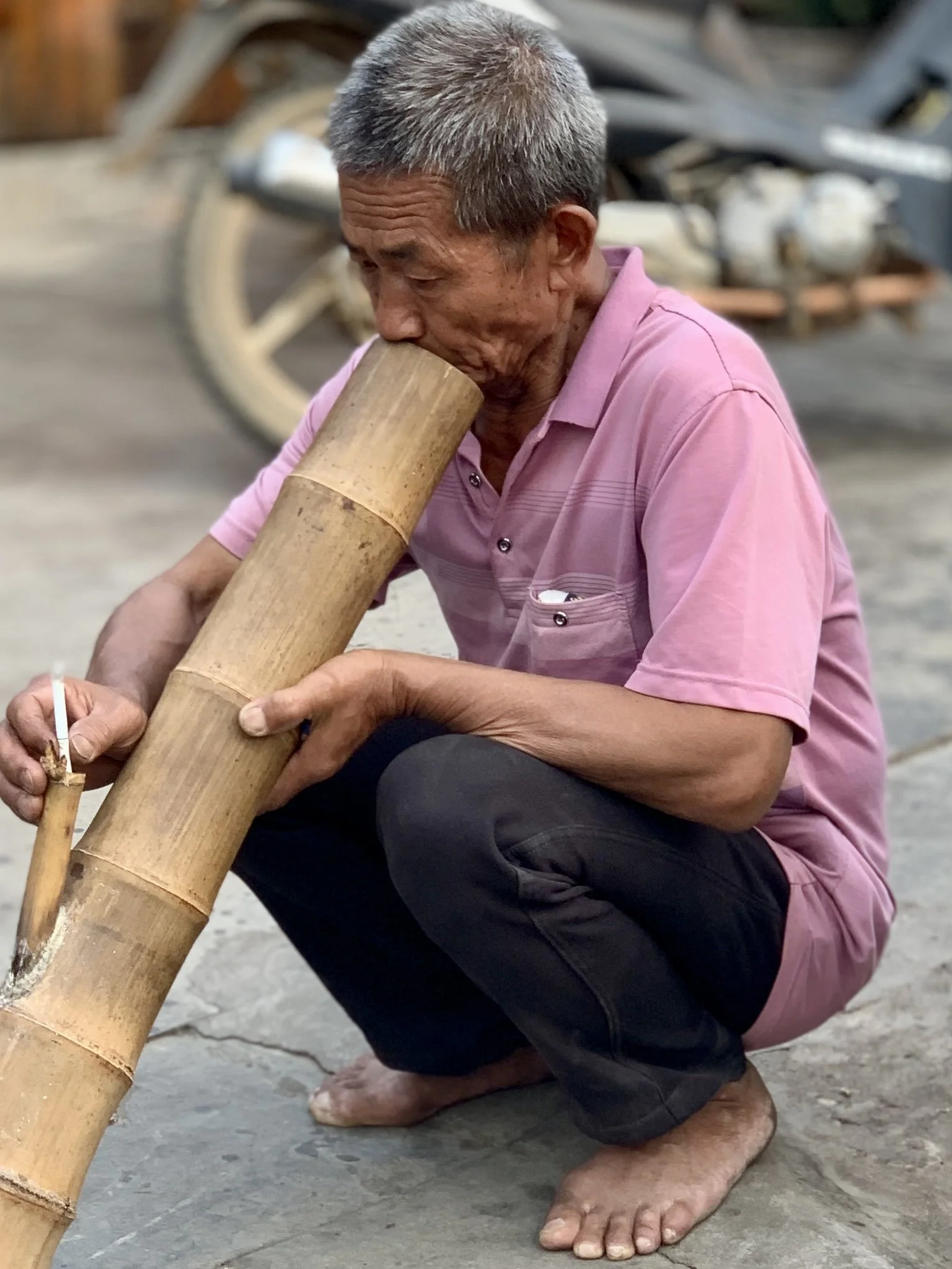An elderly man with gray hair, wearing a pink shirt and black pants, squats barefoot on the ground while blowing a fire from a bamboo pipe.