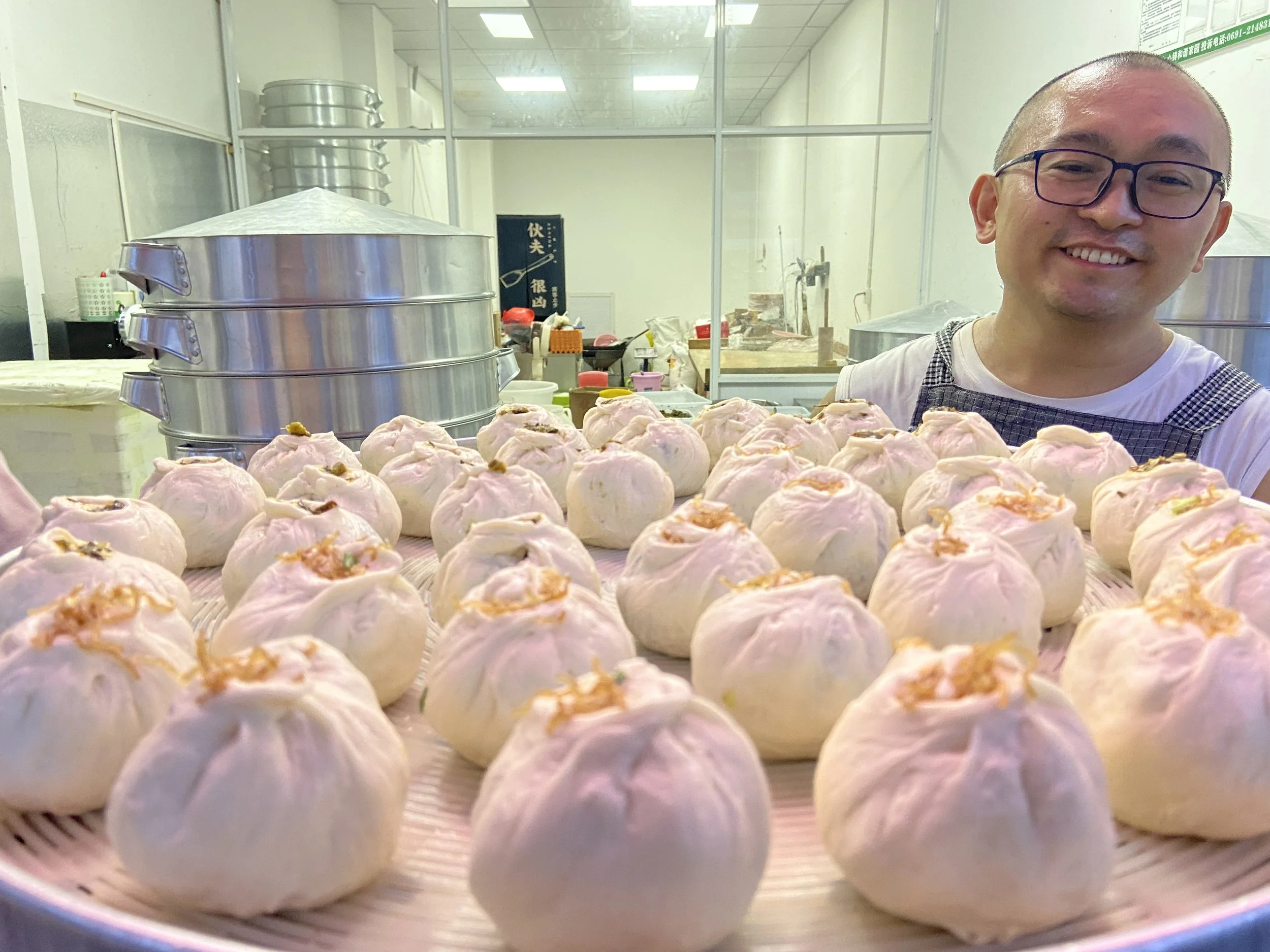 A smiling man with glasses standing behind a tray of steamed buns in a kitchen or food stall.