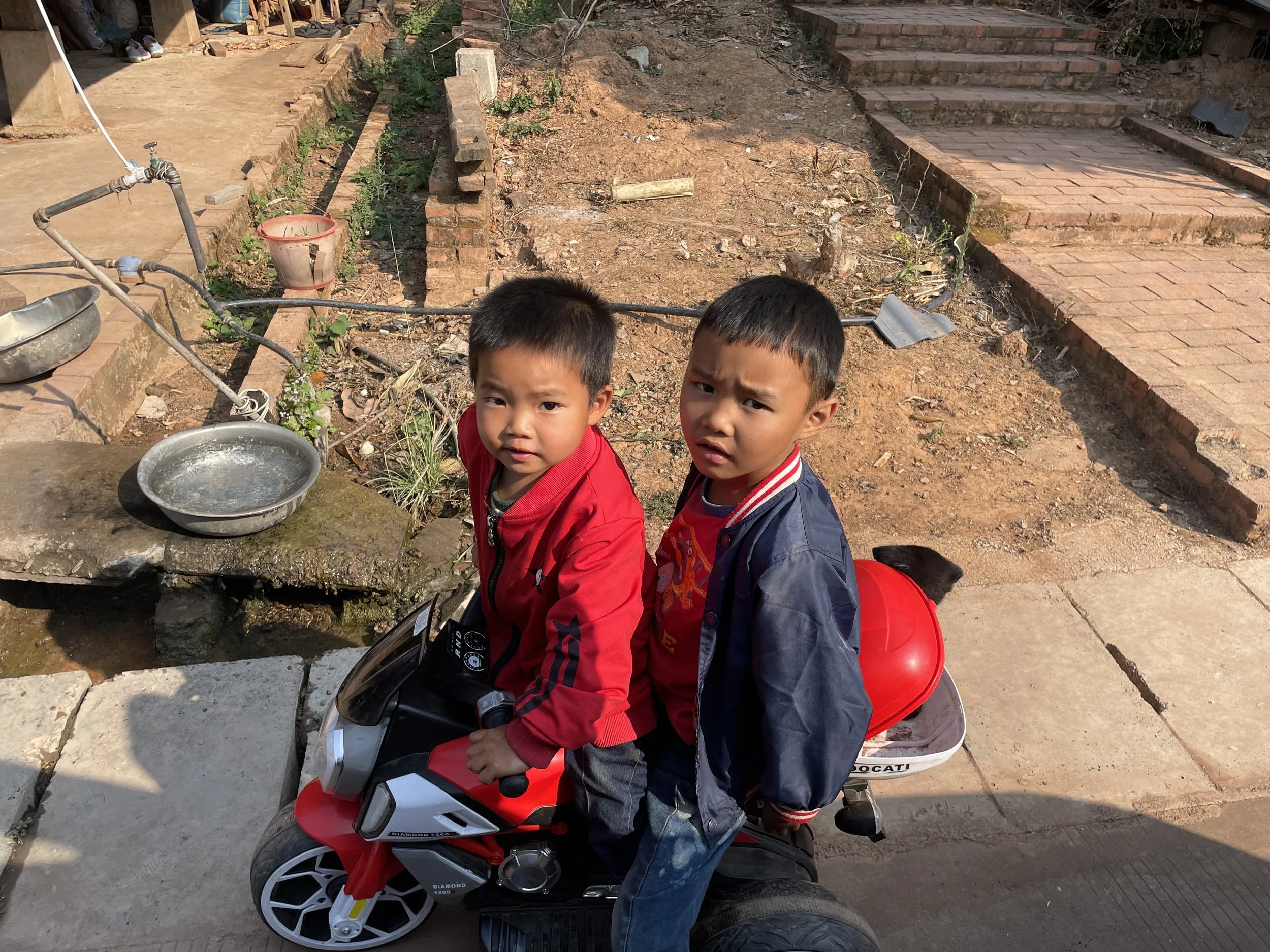 Two young boys sitting on a toy motorcycle outdoors, with one boy wearing a red jacket and the other wearing a dark blue jacket, in front of a dirt and brick area with a brick staircase and construction materials.