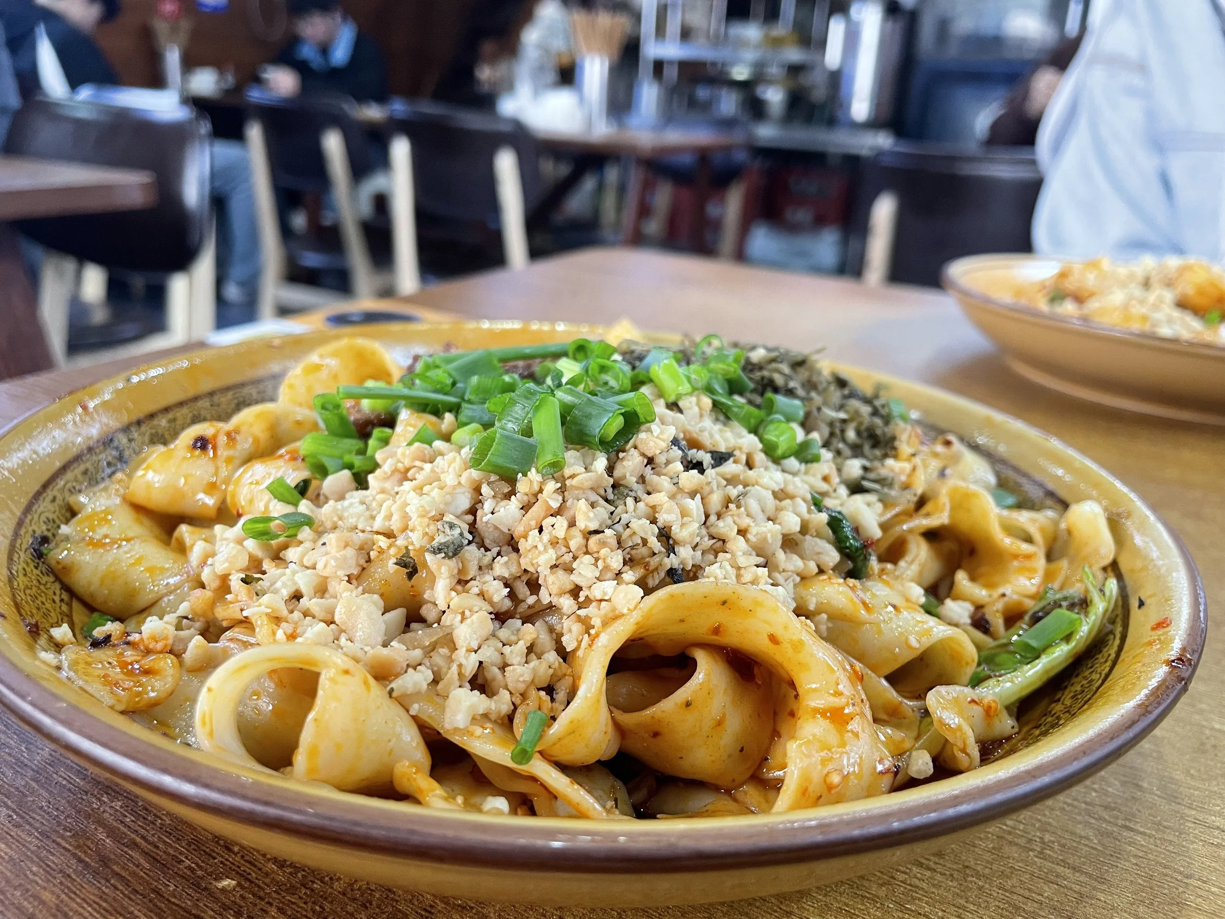 Noodle dish topped with chopped peanuts, chopped green onions, and chopped herbs in a ceramic bowl on a wooden table inside a restaurant.