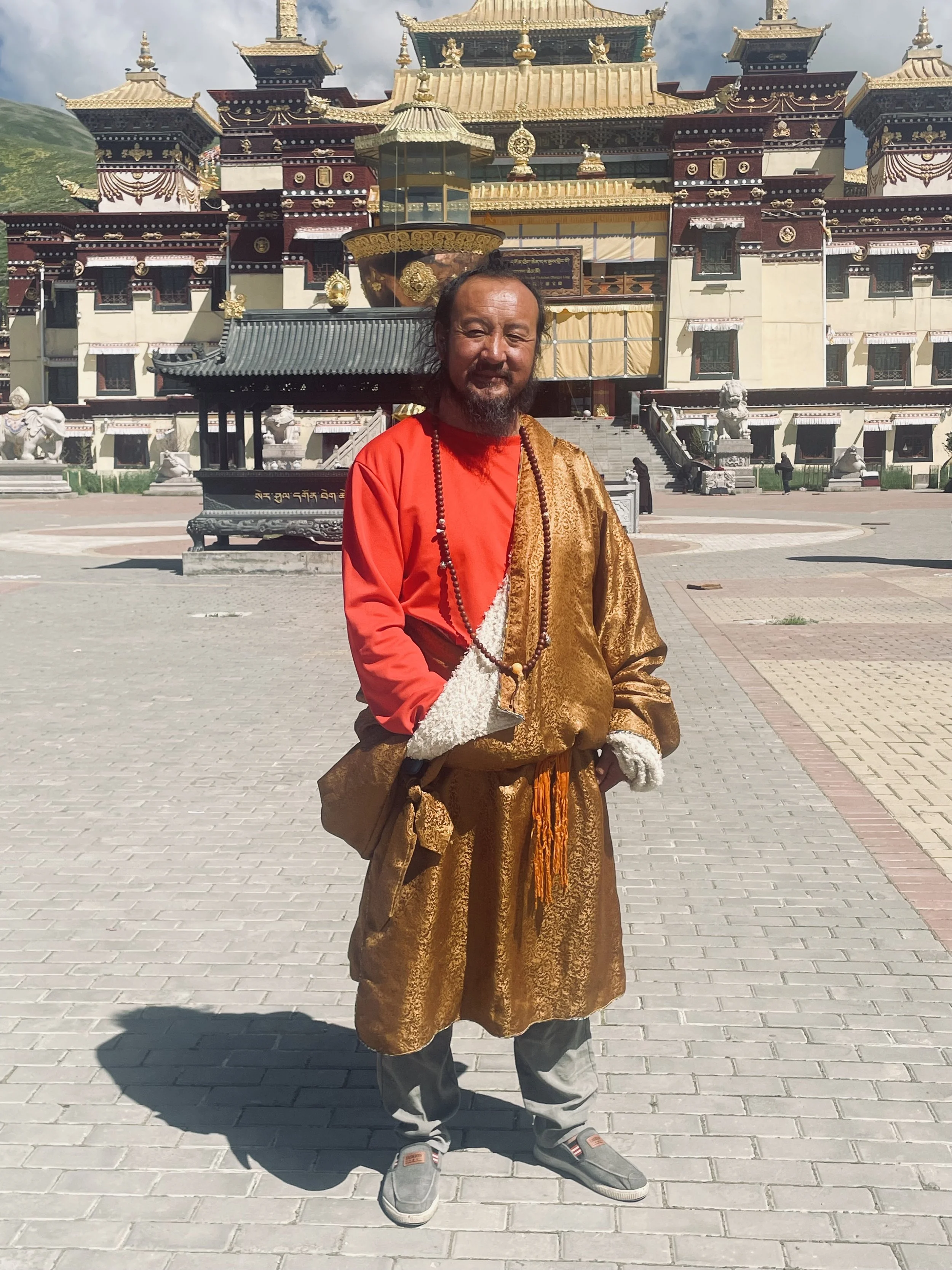 A man dressed in traditional Buddhist monk attire standing in front of a large, ornate monastery or temple with multiple roofs and intricate architectural details, under a partly cloudy sky.