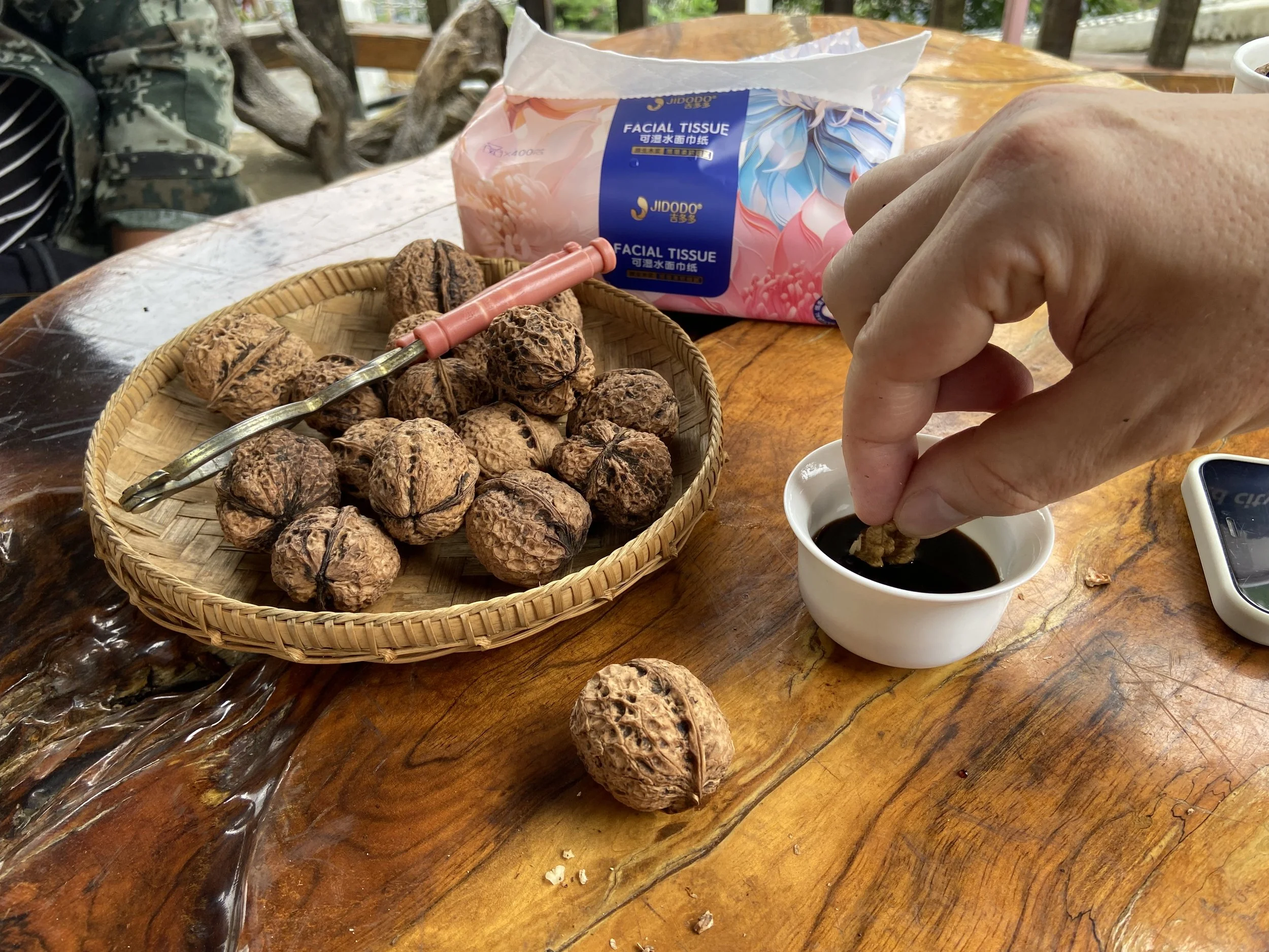 Hand dipping a walnut into a cup of dark liquid, with a basket of walnuts and a pack of facial tissue on a wooden table.
