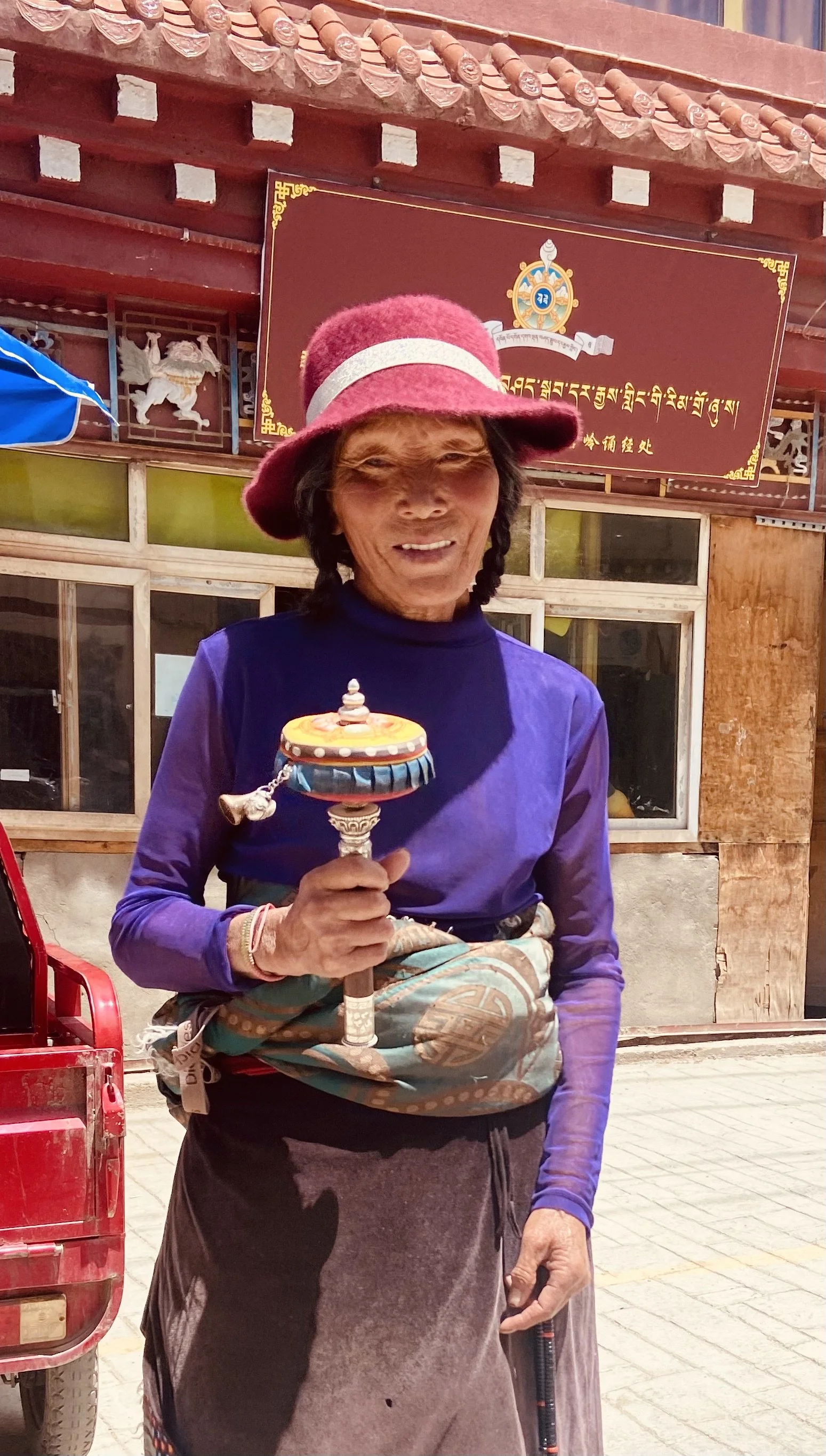 An elderly woman wearing a maroon hat, purple long-sleeve shirt, and a patterned apron holds a colorful prayer wheel in front of a building with Tibetan script and symbols.