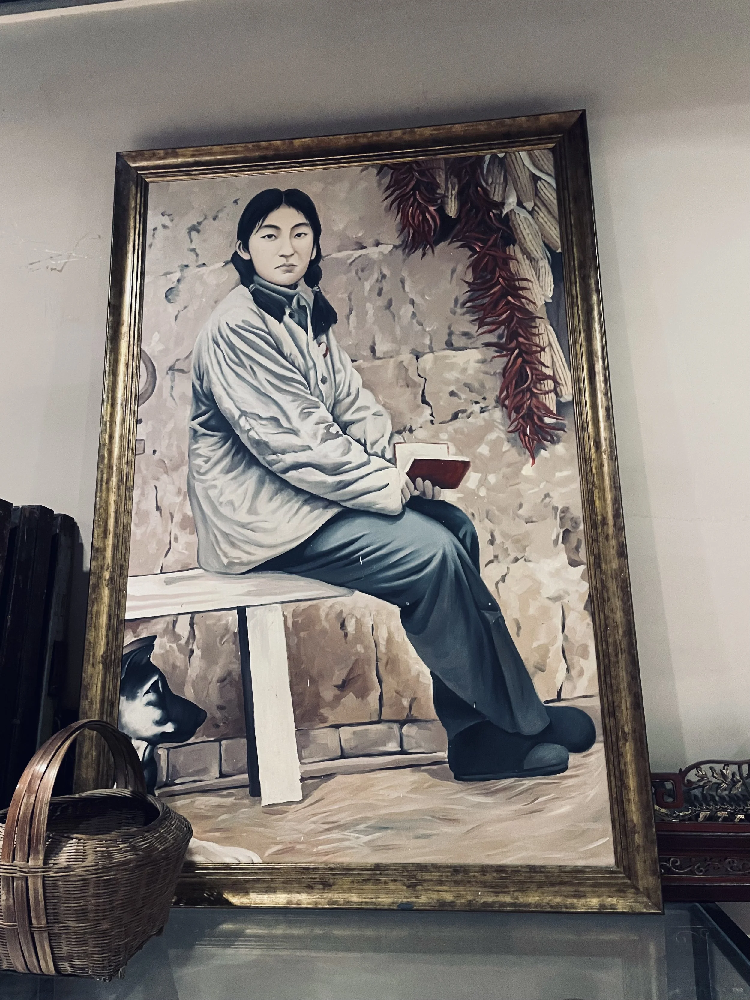 A large painting of a woman with braided hair, sitting on a wooden bench against a stone wall, holding a red book, with a dog painting at her feet, and hanging chili peppers and dried corn in the background.