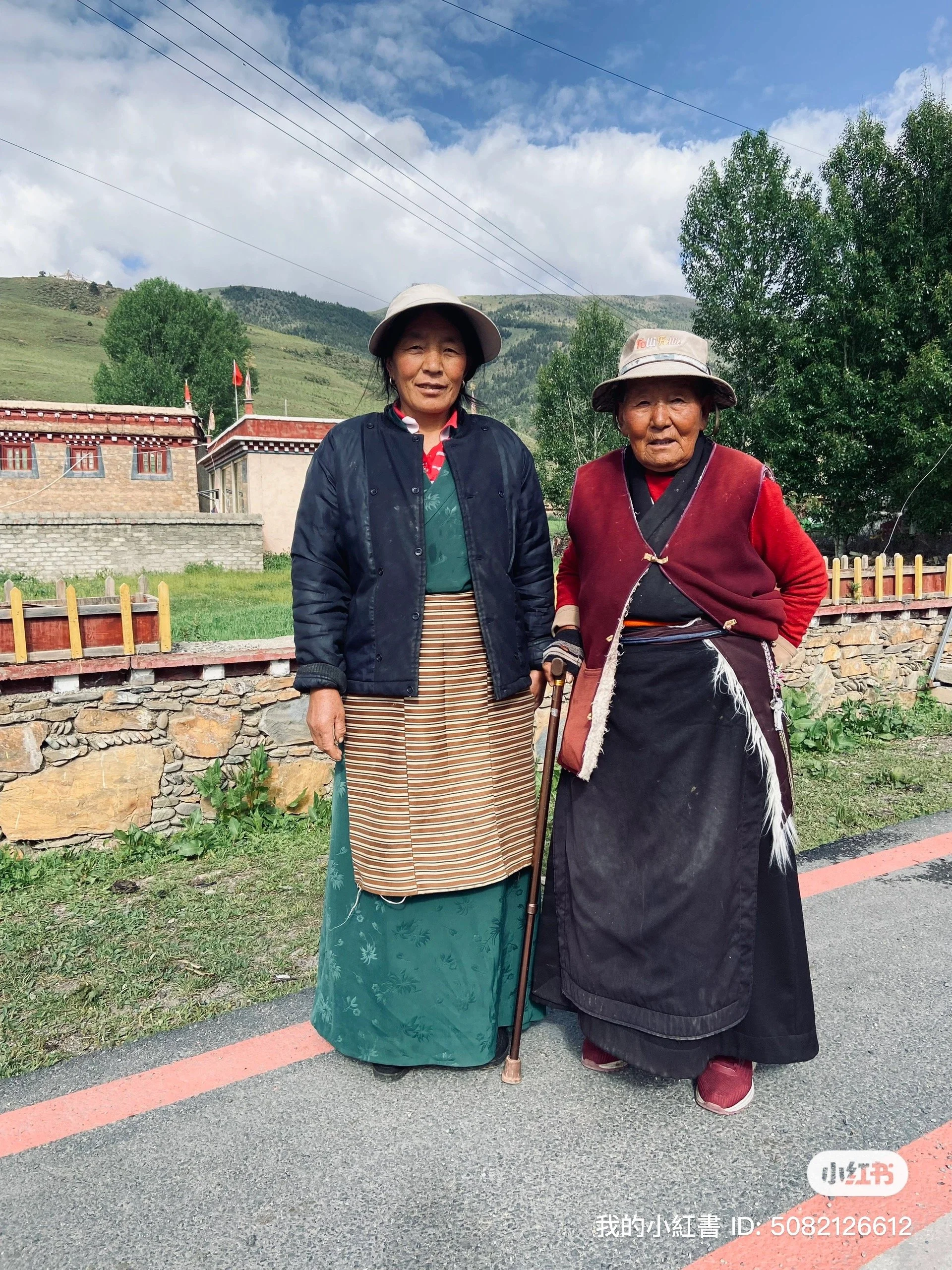 Two elderly women in traditional clothing stand outdoors on a paved road with a mountainous landscape, small building, and trees in the background.