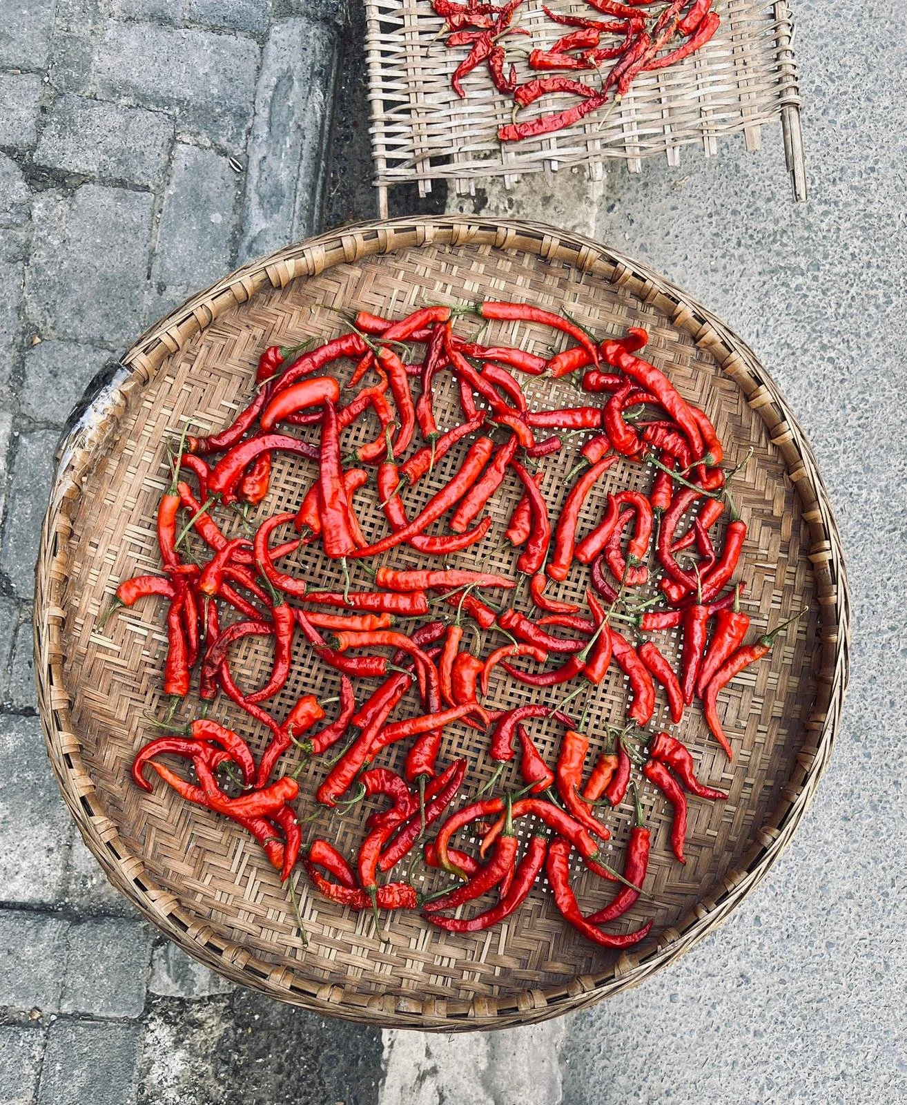 Red chili peppers drying on a woven bamboo tray placed on a stone surface.