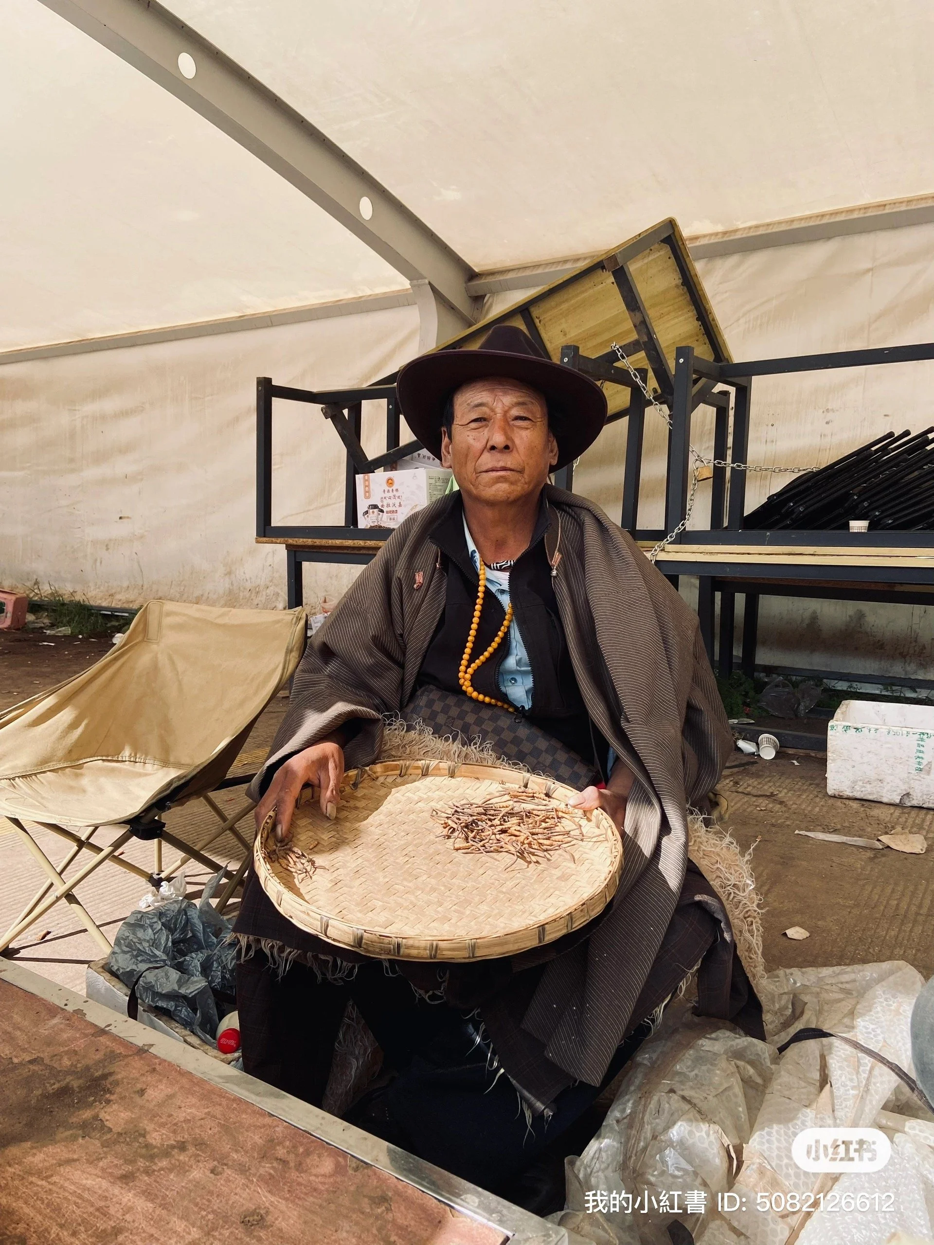 An elderly man wearing a wide-brimmed hat and traditional clothing, sitting under a tent, holding a round woven tray with dried insects.