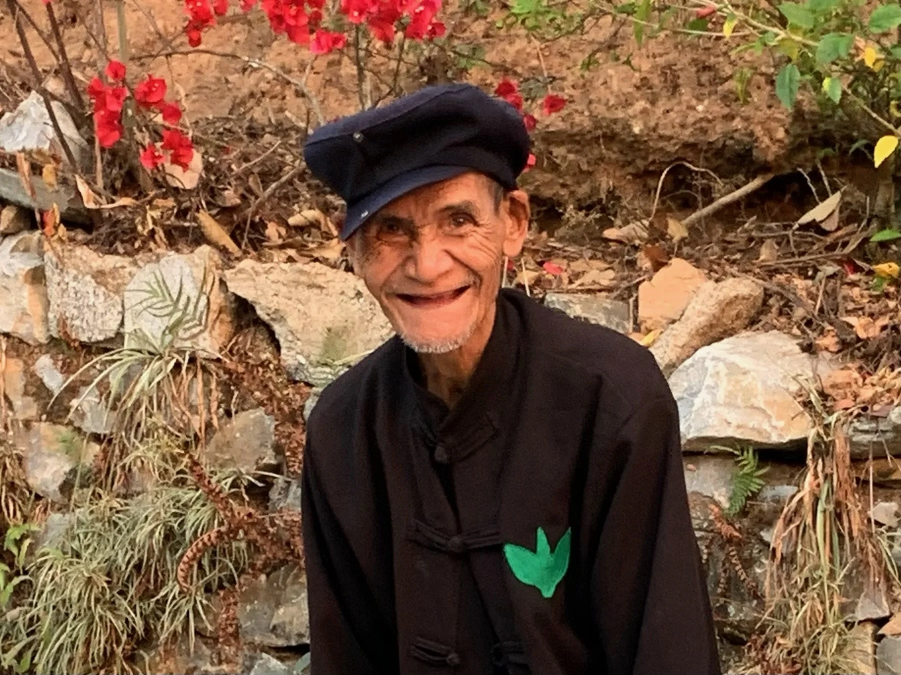 An elderly man wearing a black jacket and a black cap, smiling, standing in front of a rocky and leafy outdoor background with red flowers.