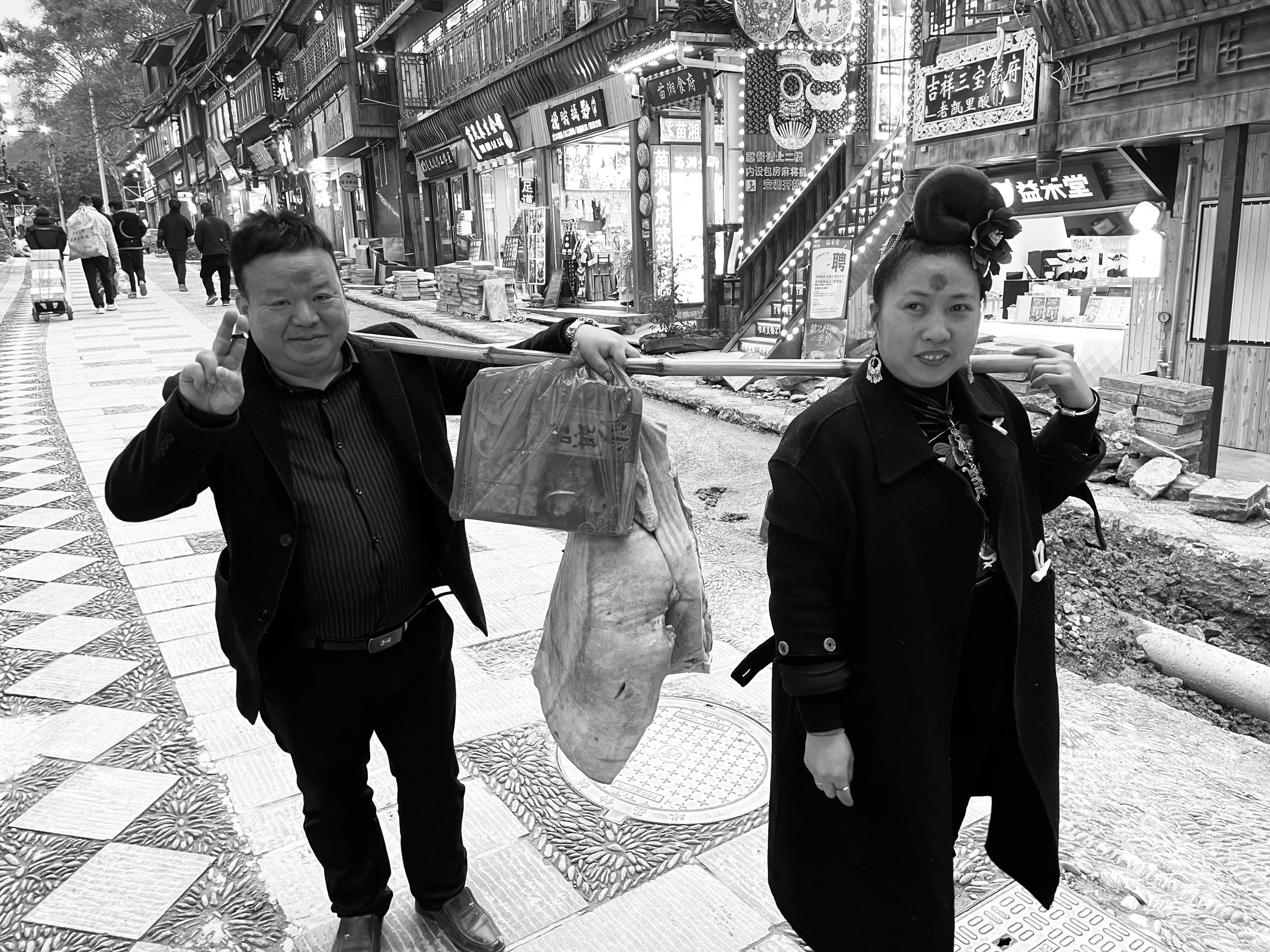 A man and a woman walking down a street, carrying goods on a pole over their shoulders, with shop signs and people in the background.