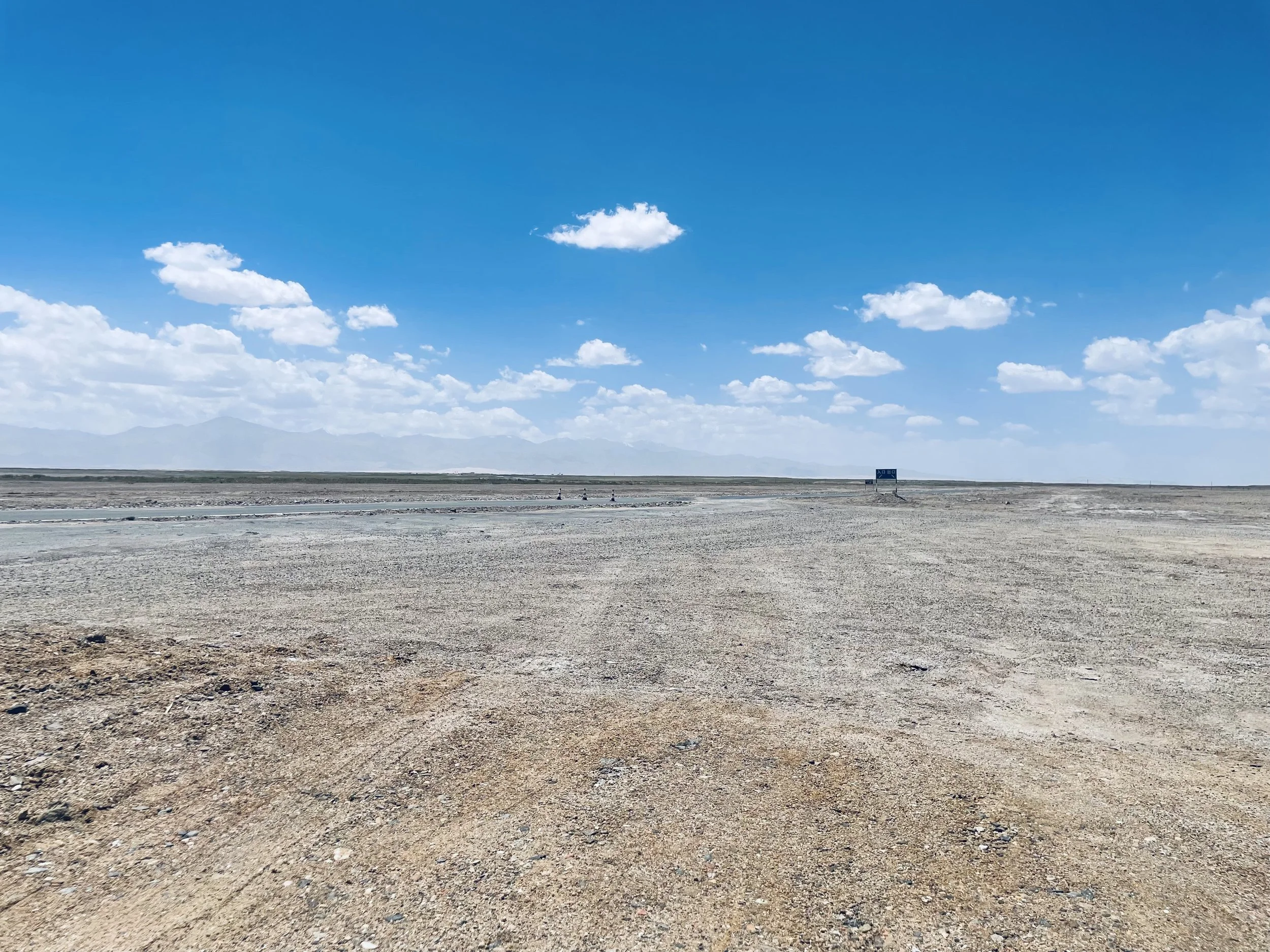 A vast, open desert landscape under a blue sky with scattered white clouds, featuring a small sign in the distance.