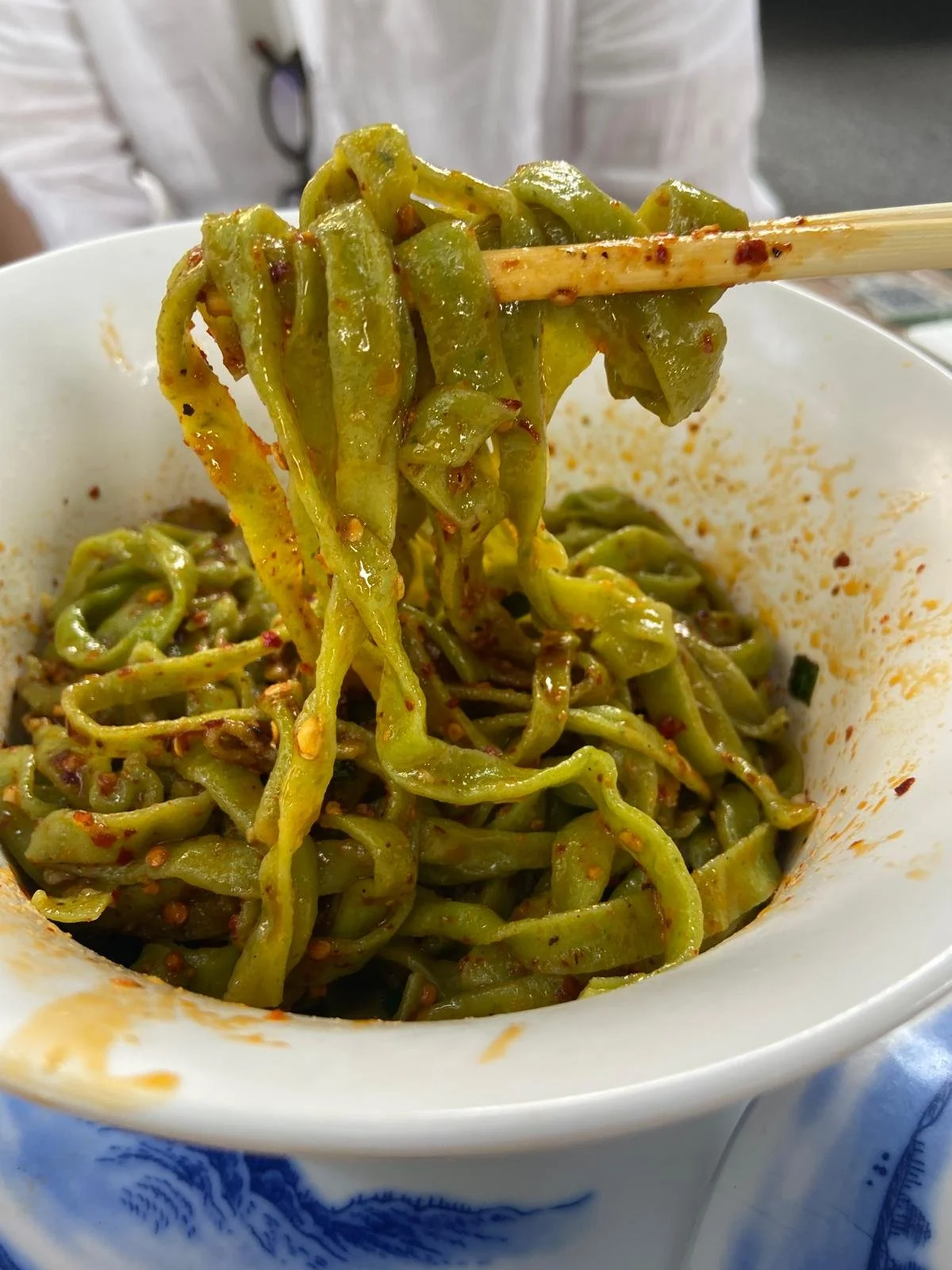 Close-up of a bowl of spicy green Asian noodles with chili flakes, with chopsticks lifting some noodles.