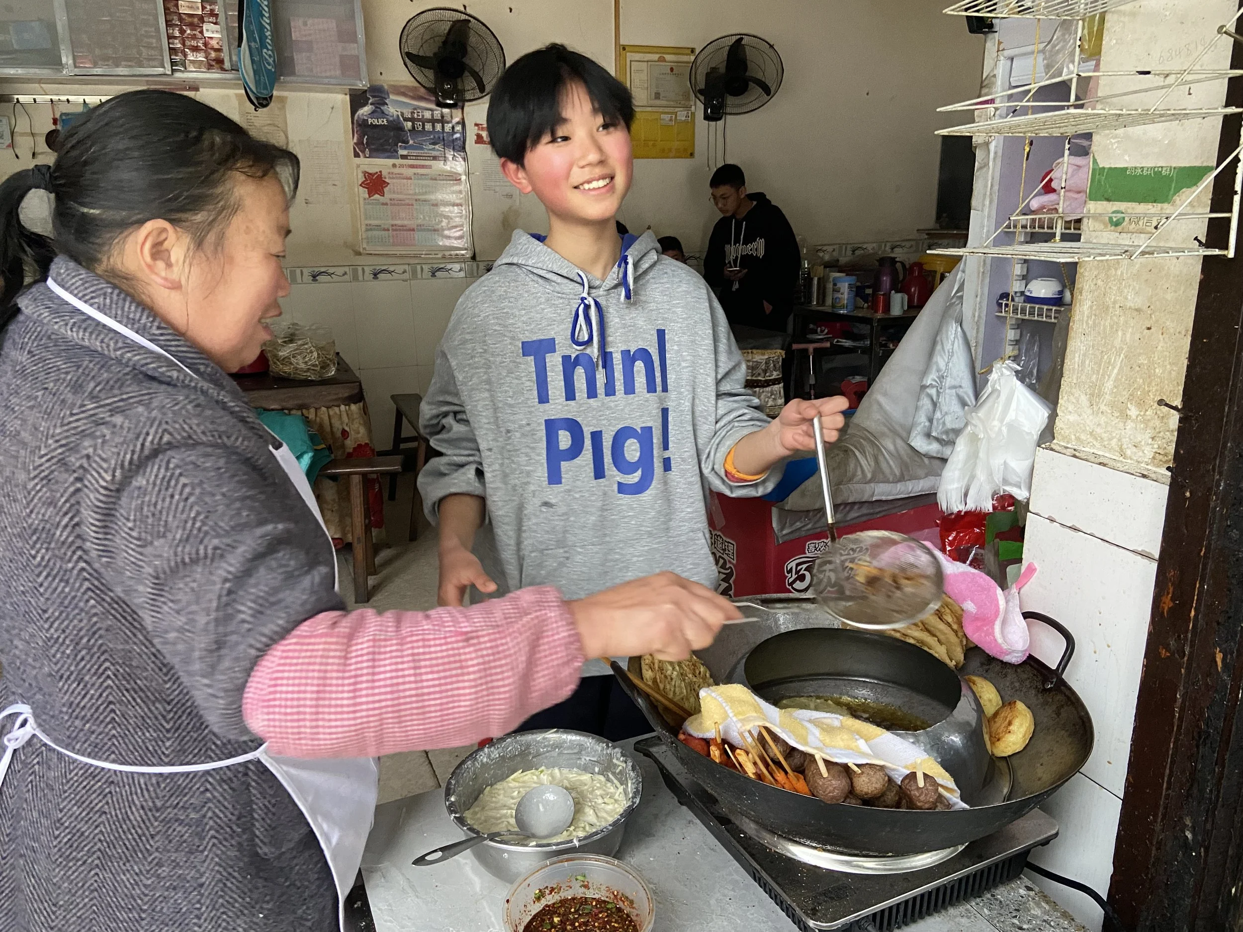 Two women cooking together at a small street food stall, cooking meatballs and vegetables in a wok, with various condiments and utensils on the counter.