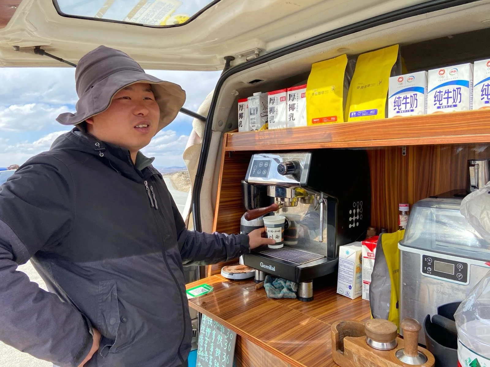 Man wearing a black jacket and wide-brimmed hat making coffee at a mobile coffee station outdoors.