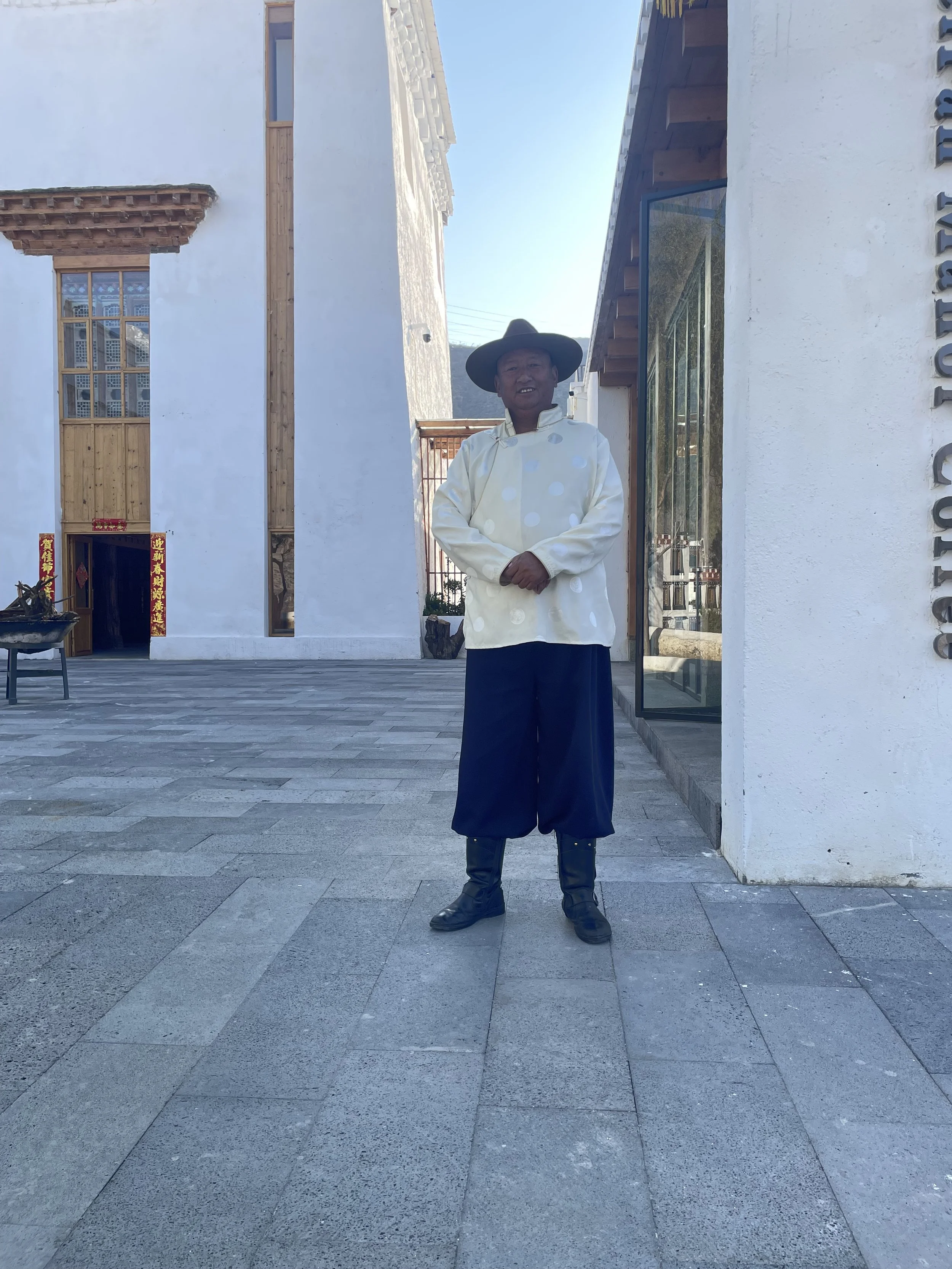 A person dressed in traditional or historical attire, wearing a wide-brimmed hat, a cream-colored coat with polka dots, dark wide-leg pants, and black boots, standing outside in front of a modern white building during daylight.