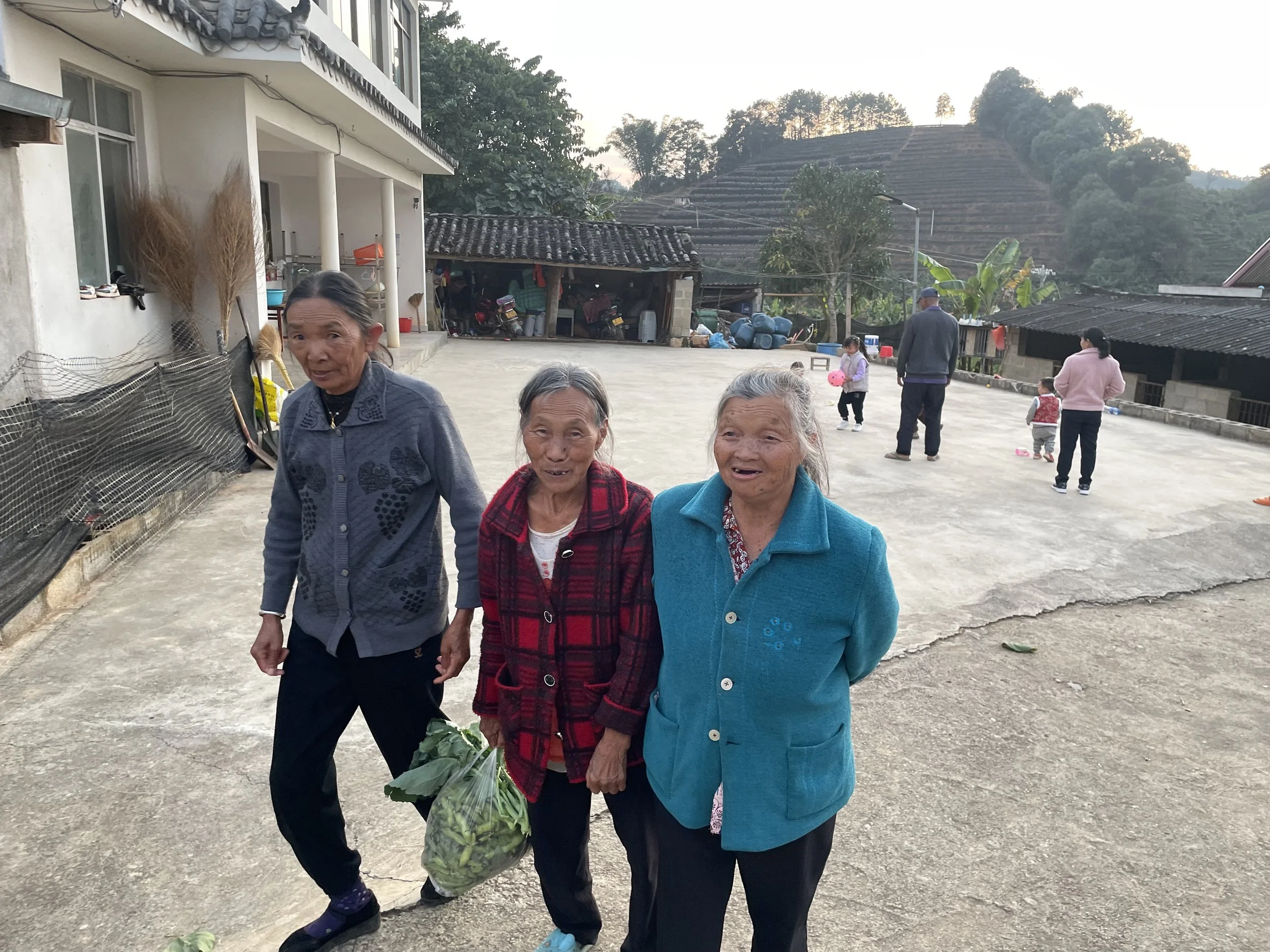 Three elderly women standing outside on a concrete area, with two women holding a plastic bag of vegetables, in a rural village setting with houses and children playing in the background.
