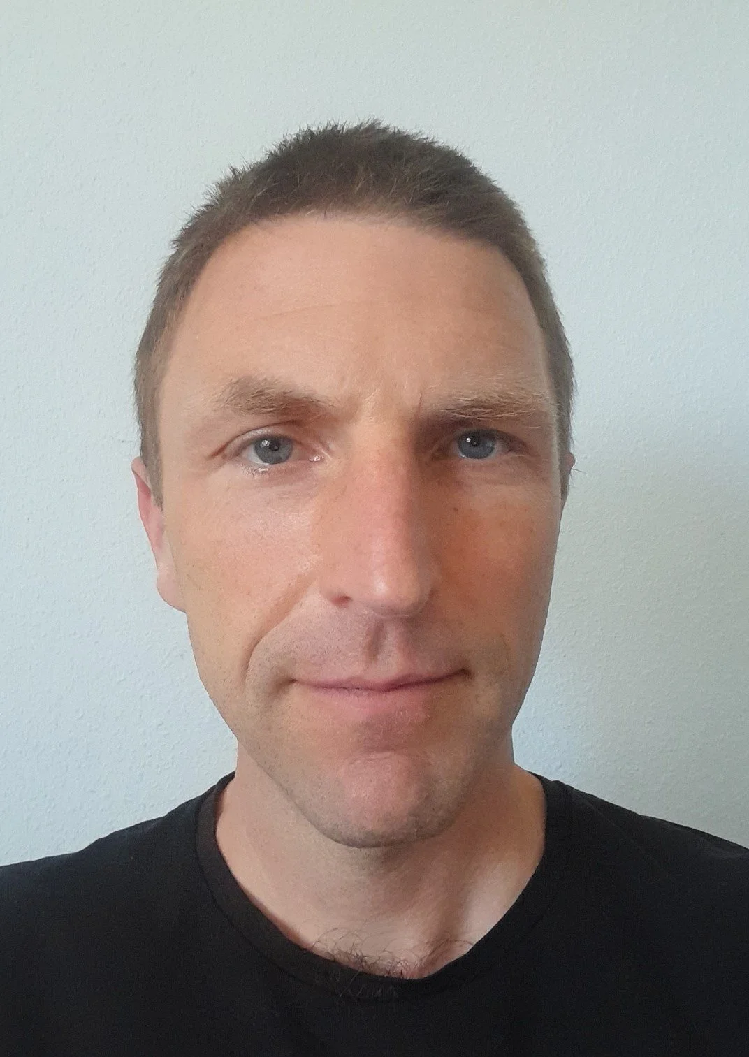 A close-up portrait of a man with short brown hair, blue eyes, and fair skin, wearing a black T-shirt, standing in front of a plain light-colored wall.