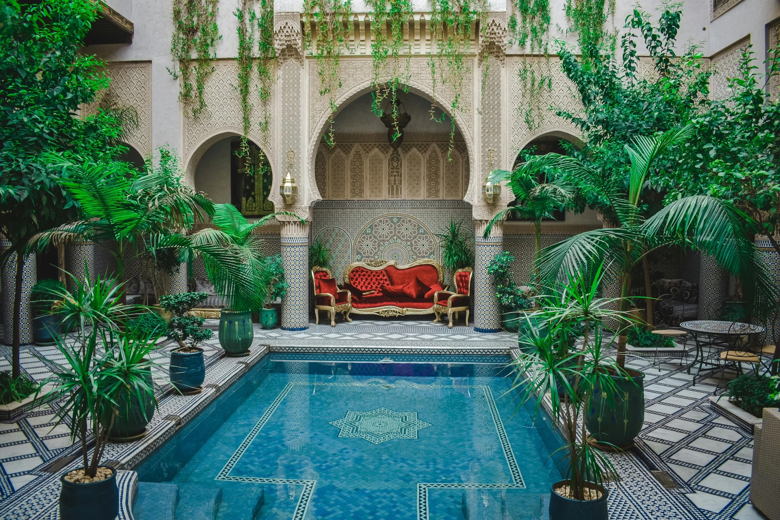Traditional Moroccan riad courtyard with ornate arches, mosaic-tiled pool, lush greenery, and red velvet seating.