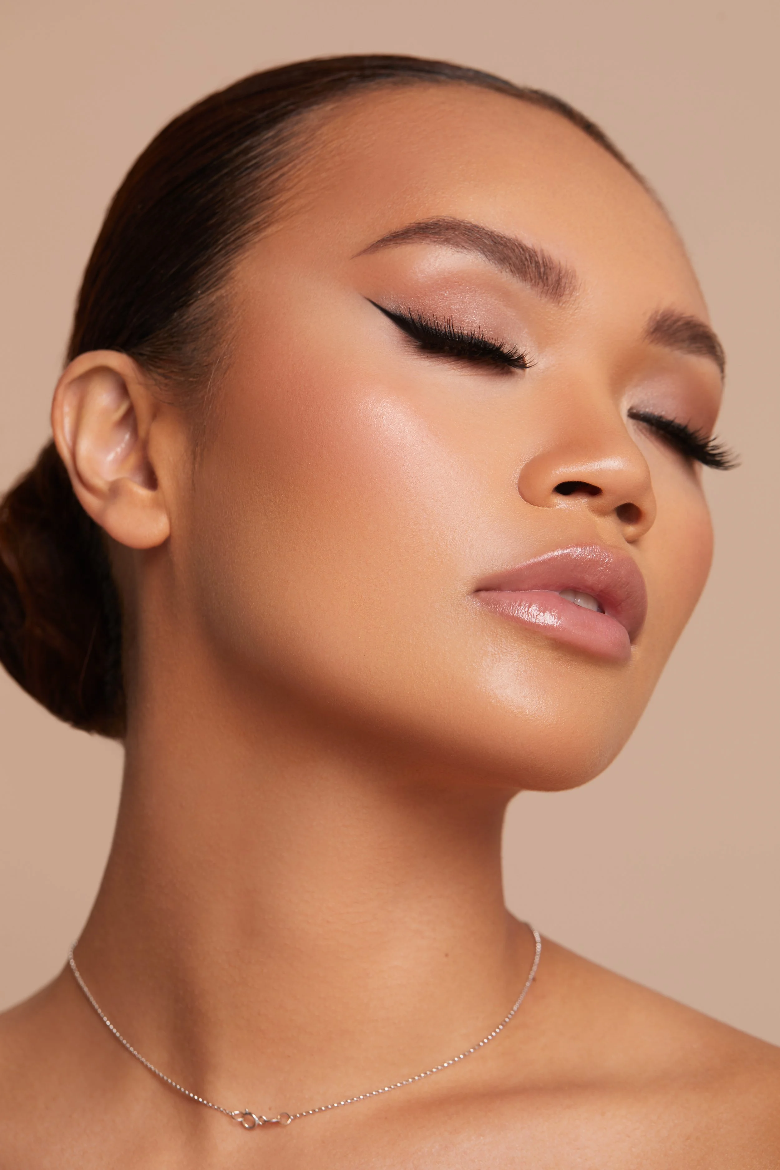 Special occasion hair and makeup on Asian lady in Sydney featuring winged eyeliner, glowing skin, glossy nude lips and a sleek low bun.
