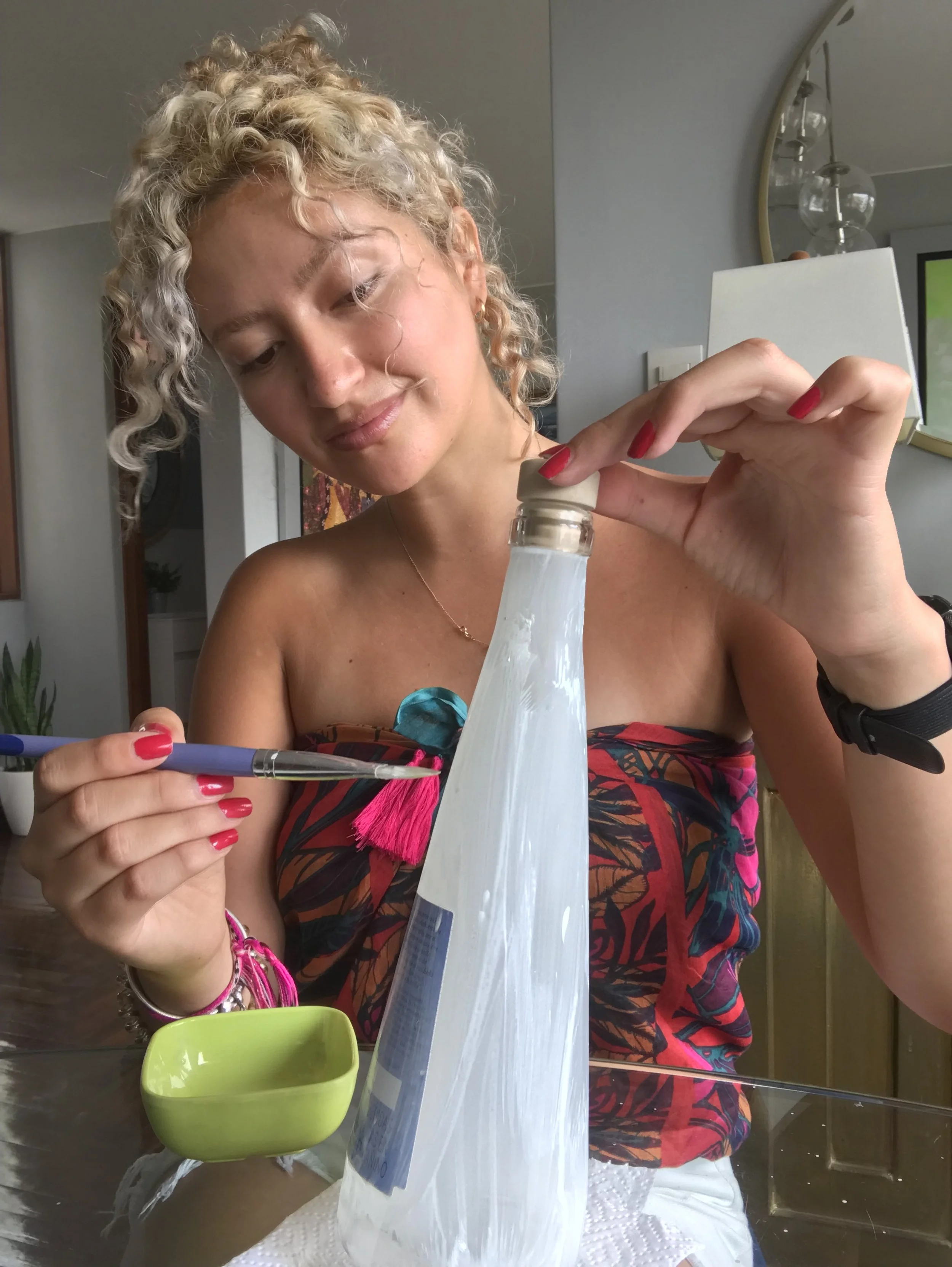 Woman with curly blonde hair sitting at a table, signing a glass bottle with a pen, wearing a colorful strapless top and pink nail polish.