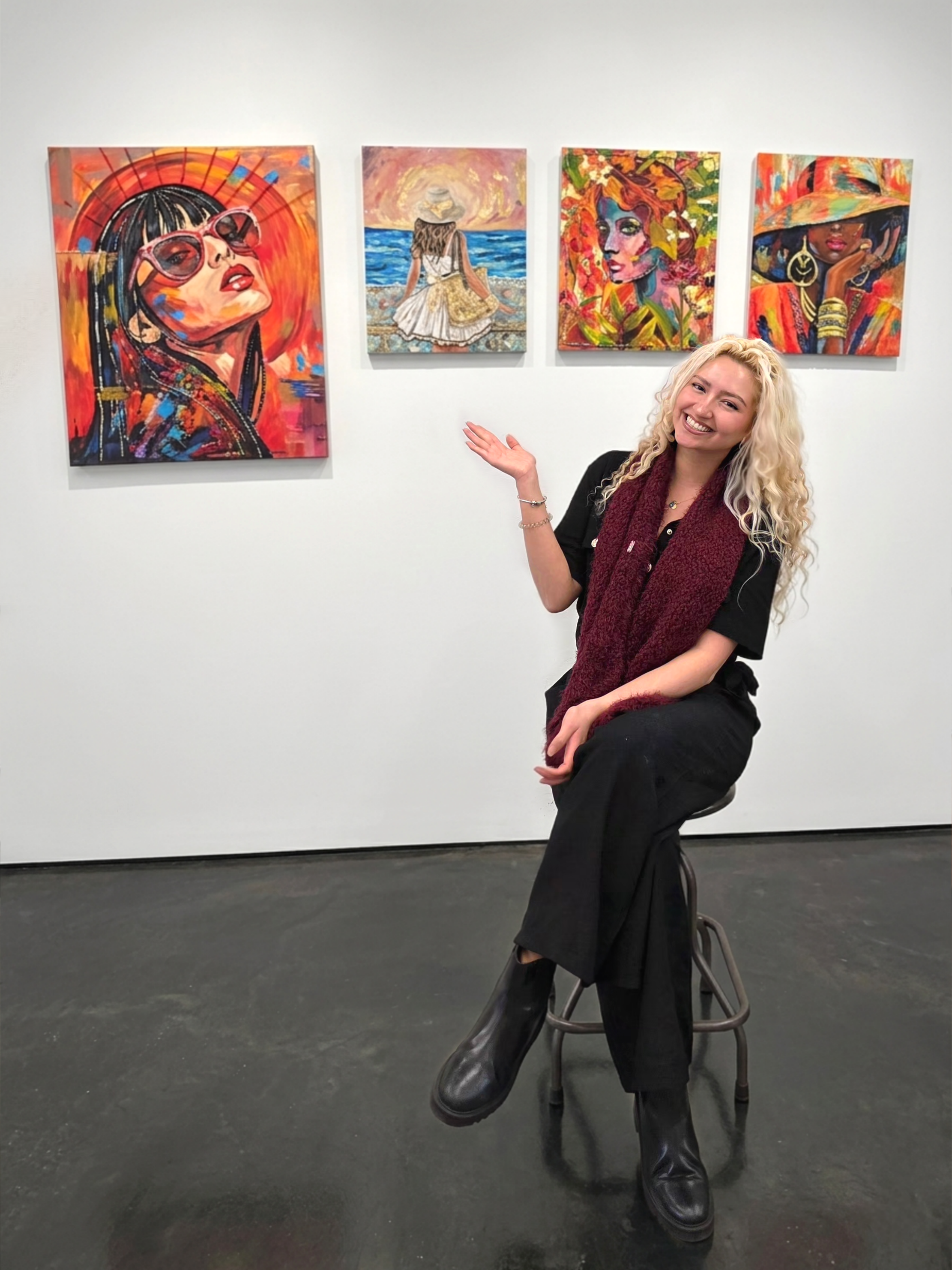 A woman with curly blonde hair and black clothing sitting on a stool in an art gallery, pointing at four vibrant paintings of women wearing colorful outfits and accessories on a white wall.
