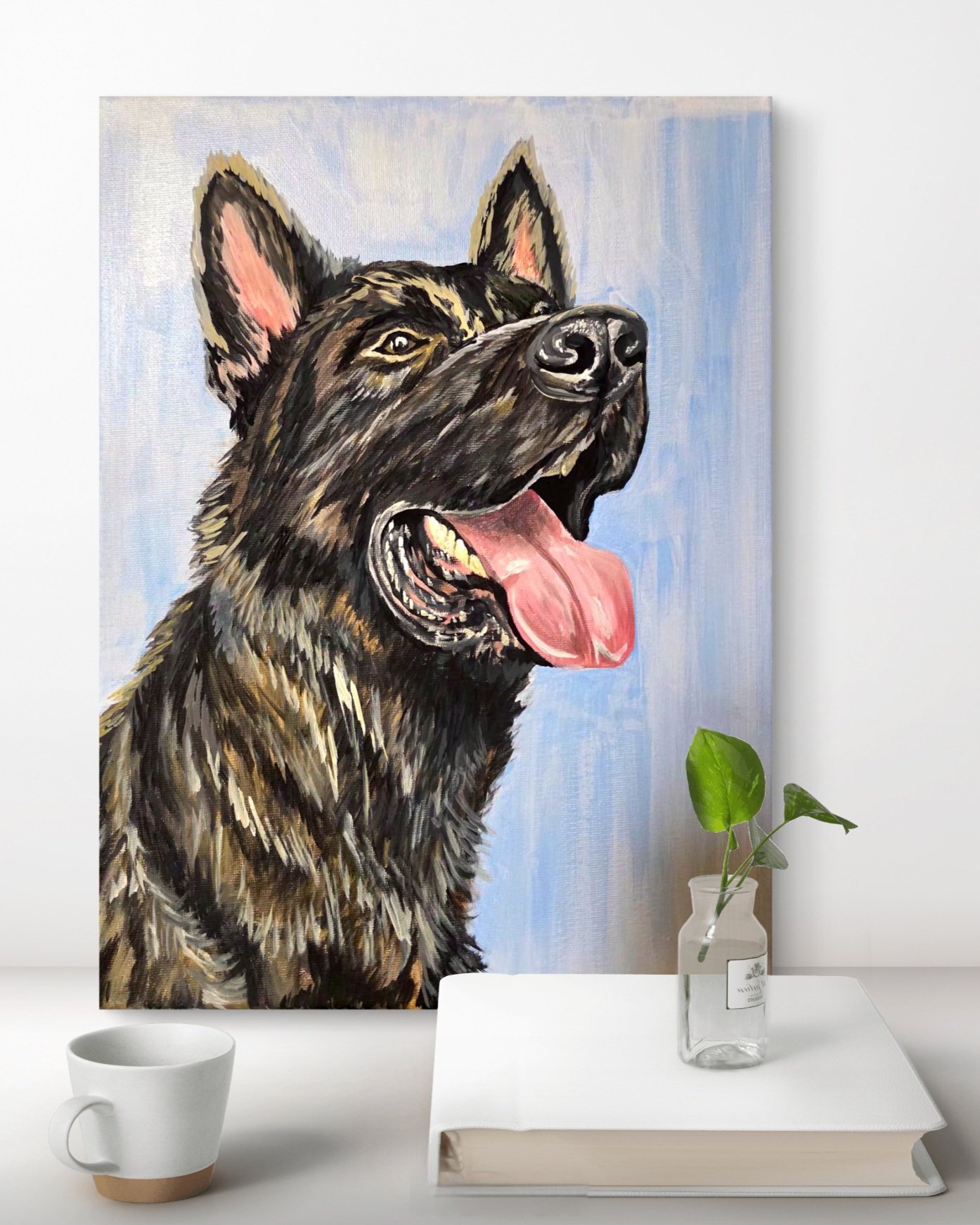 A painting of a German Shepherd dog with its tongue out, hanging on a wall behind a white table. The table has a glass vase with green leaves, a closed book, and a white mug.