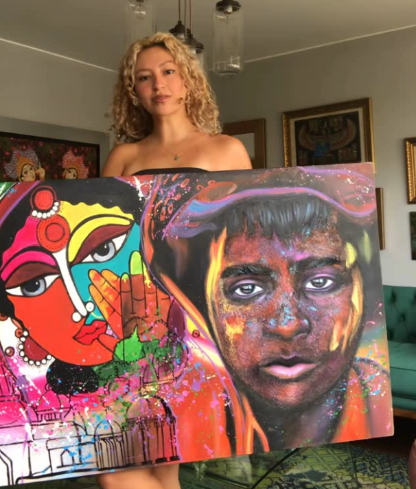 Young woman with curly blonde hair holding a large colorful painting. The artwork features a stylized female face with vibrant colors and abstract patterns, alongside a realistic portrait of a young man with a painted face and a hat.