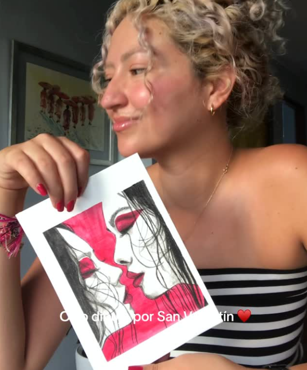 A woman with curly blonde hair and tan skin, smiling and looking at a drawing she is holding. She is wearing a black and white striped off-the-shoulder top, small hoop earrings, a thin necklace, and red nail polish. The drawing features two women facing each other with red lips and hair, in a stylized black, white, and red design. In the background, there is a framed artwork on the wall.