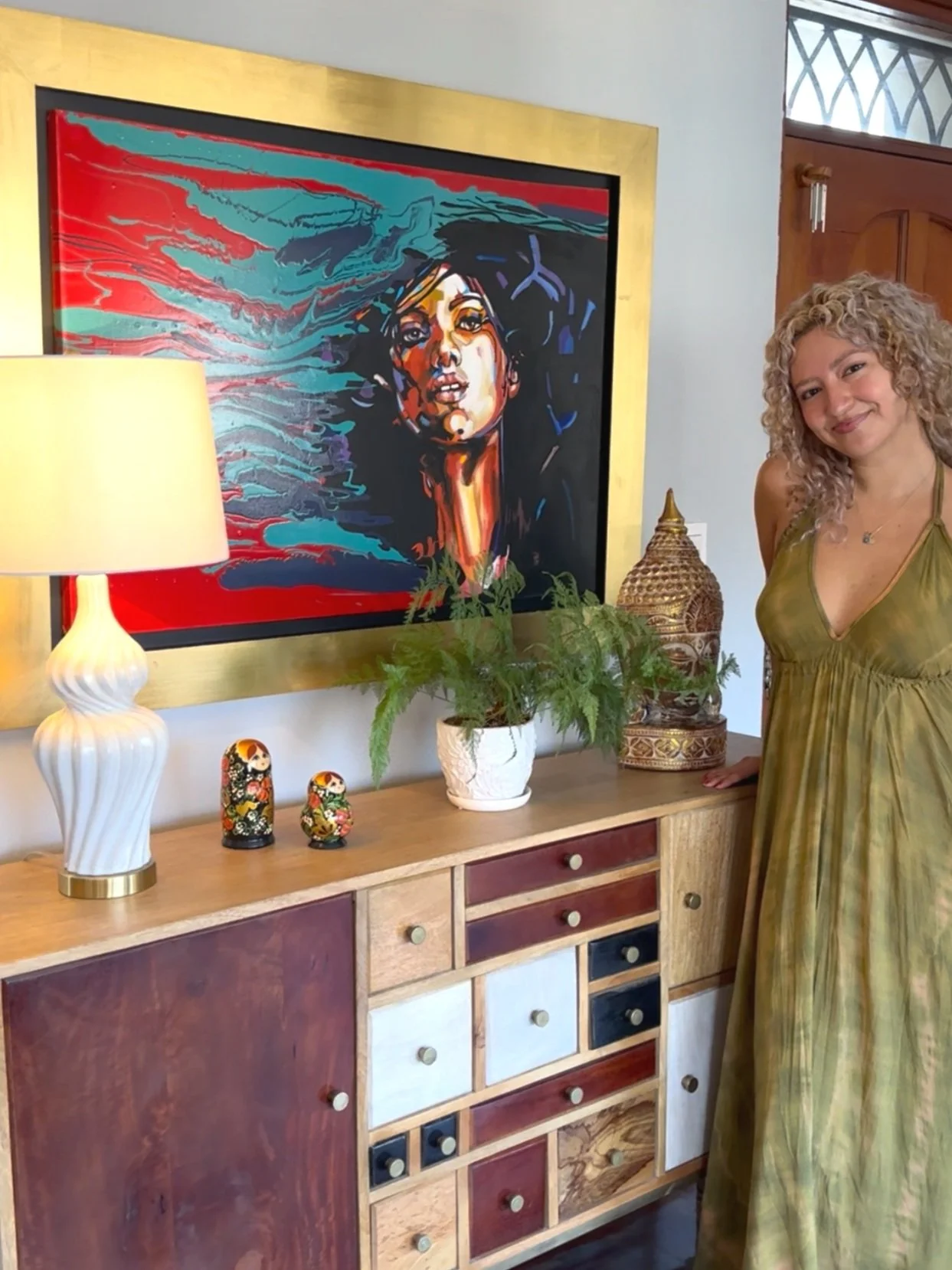 A woman with curly blonde hair wearing a green dress standing next to a wooden sideboard with multi-colored drawers. On the wall above, there's a large framed painting of a woman with flowing hair, painted in vibrant colors. The sideboard holds a white lamp, two Russian nesting dolls, a white pot with green ferns, and a decorative jar.