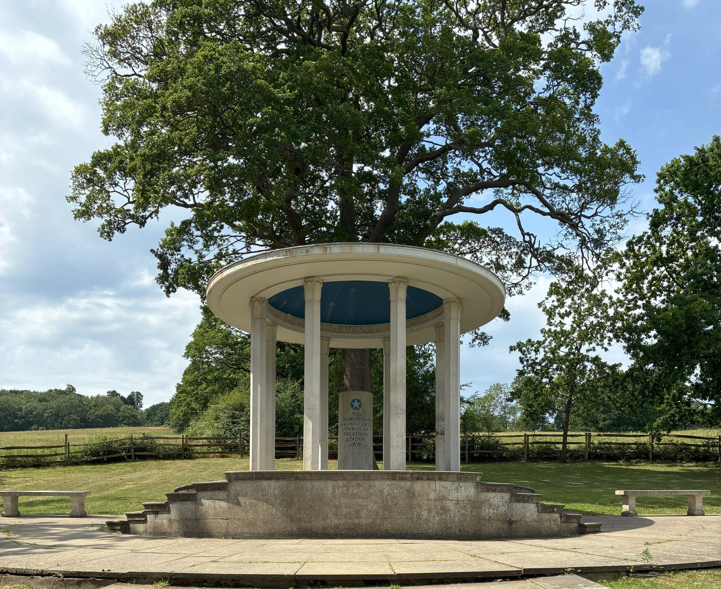 A white, circular bandstand with a domed roof and columns, situated in front of a large deciduous tree with green leaves. There are steps leading up to the bandstand and a stone plaque with an inscription. The background shows a field and more trees, with a partly cloudy sky overhead.
