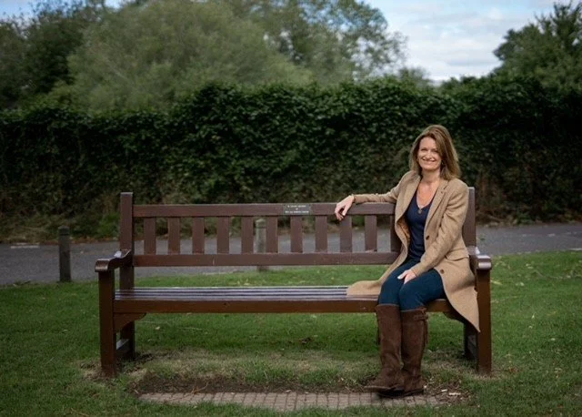 A smiling woman sitting on a wooden park bench outdoors with greenery and trees in the background.