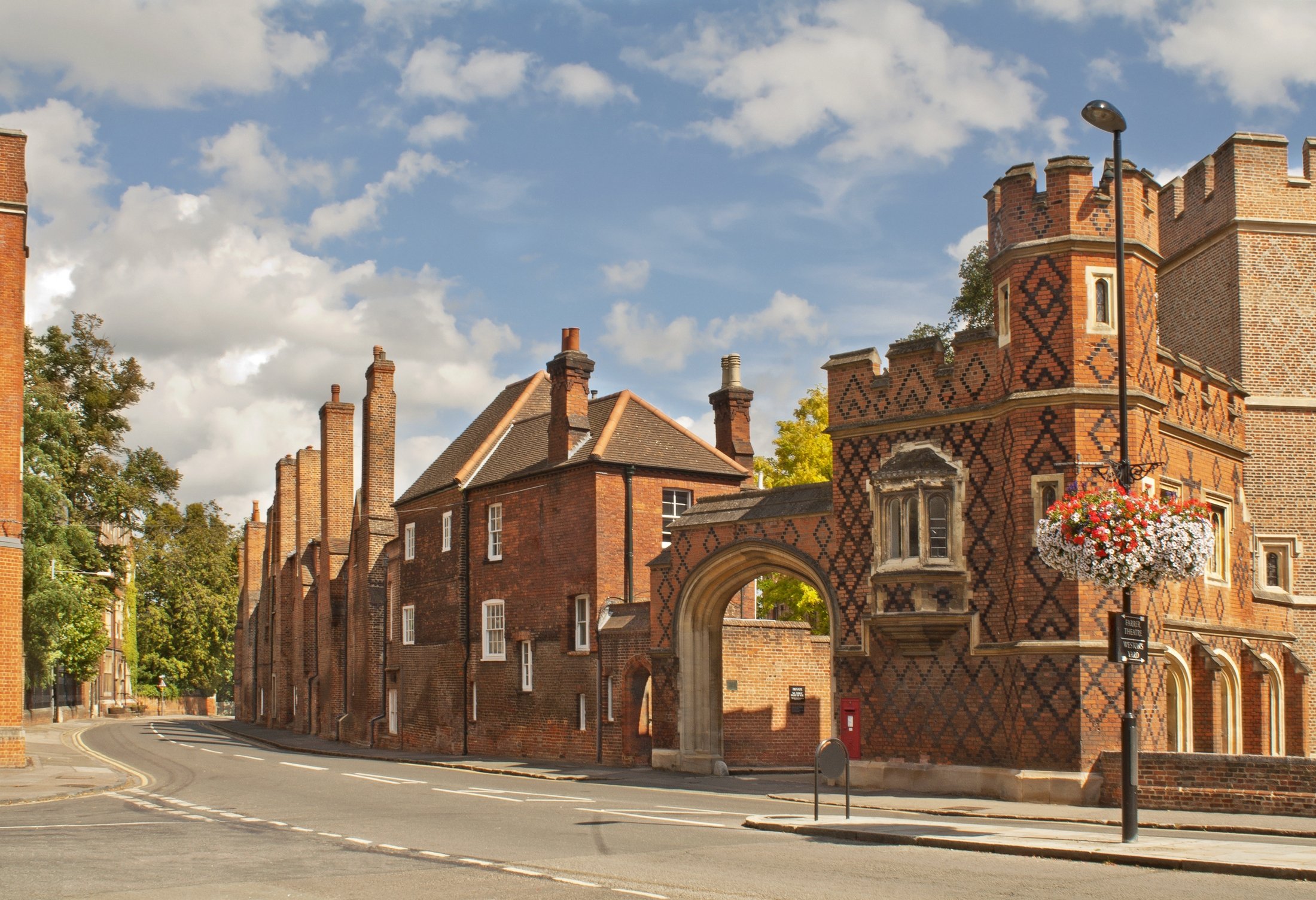 A street scene with historic red brick buildings featuring medieval architecture, including turrets and archways, under a partly cloudy sky, with a flower basket hanging outside one building and street lighting posts visible.