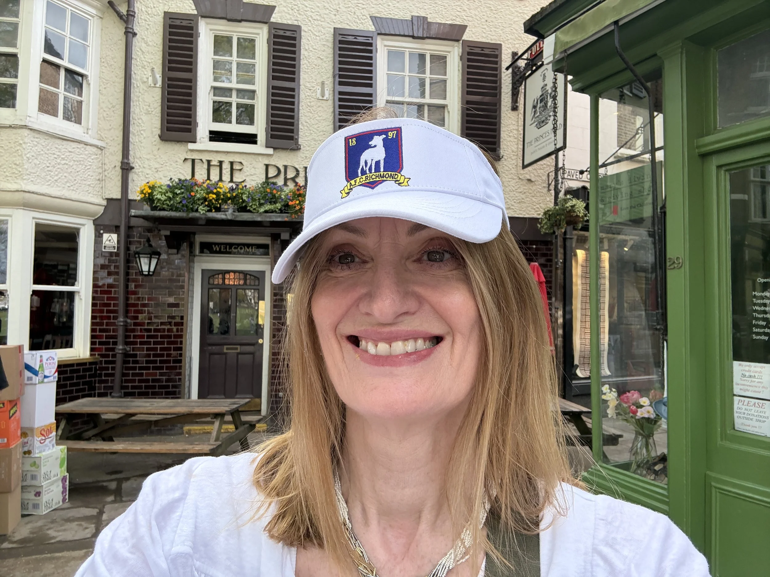 Smiling woman wearing a Ted Lasso visor, taking a selfie outside The Princes Head Pub Richmond.