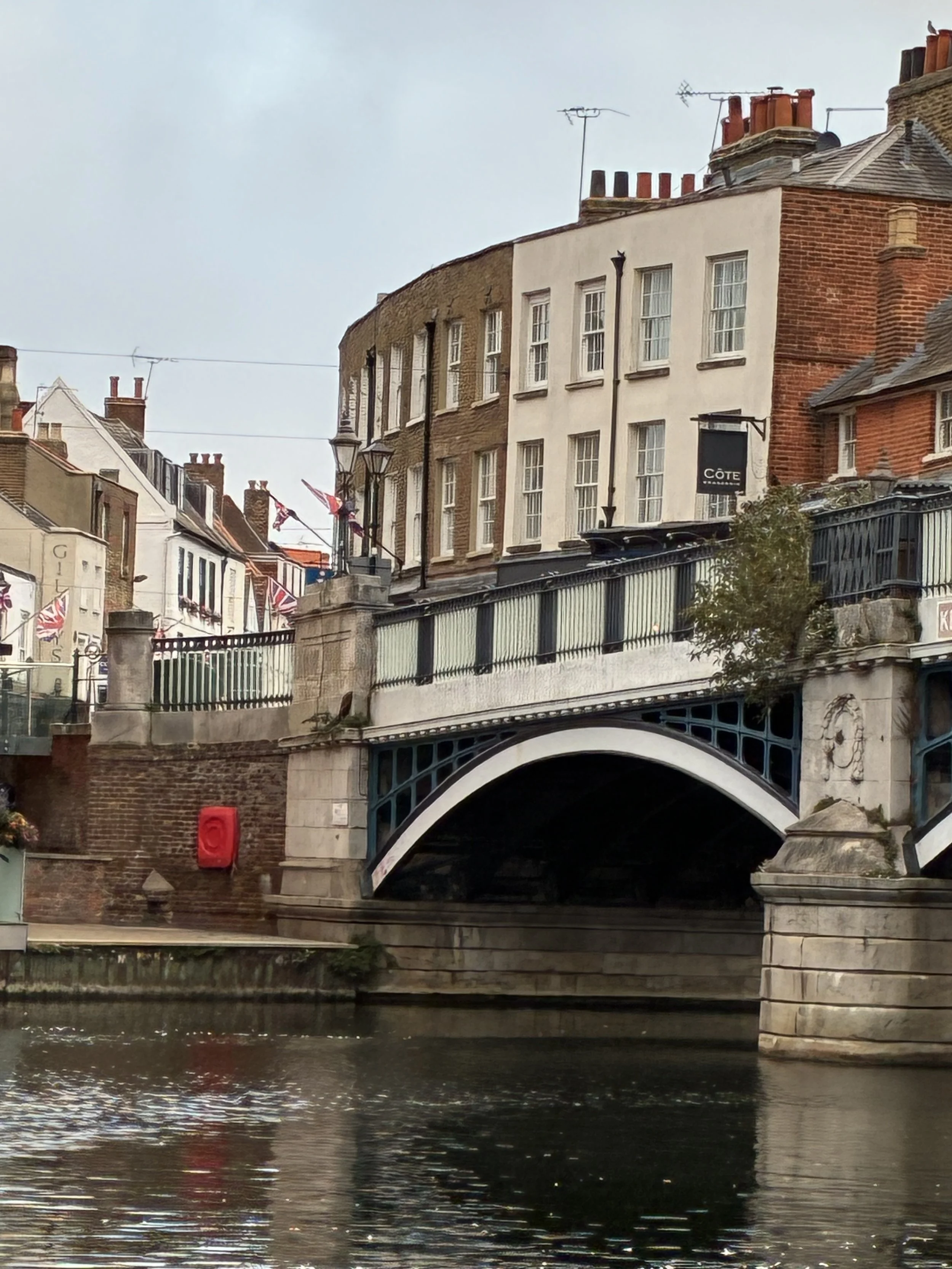 A canal with a bridge passing over it, lined with buildings featuring brick and painted exteriors, some with flags and multiple chimneys on the rooftops.